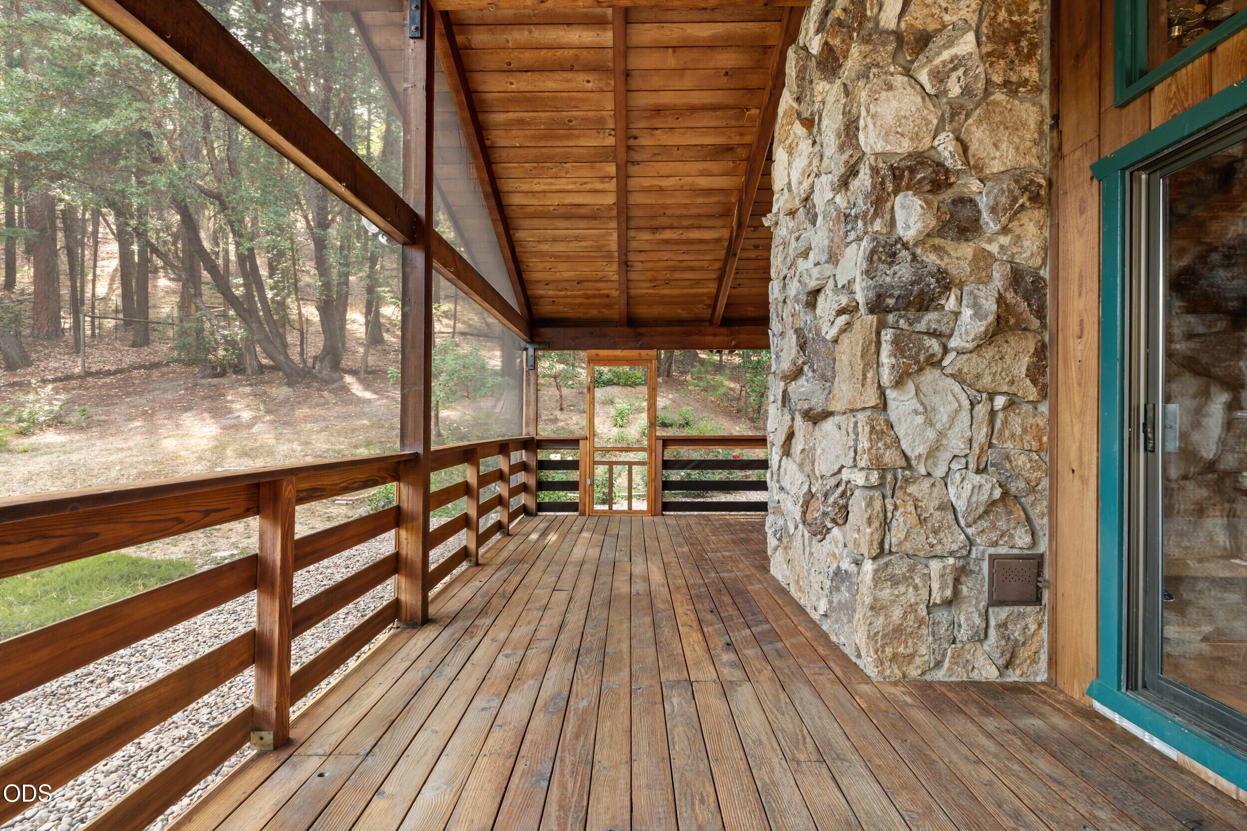 Covered wooden porch with stone wall, wood railings, and a view of the wooded yard.