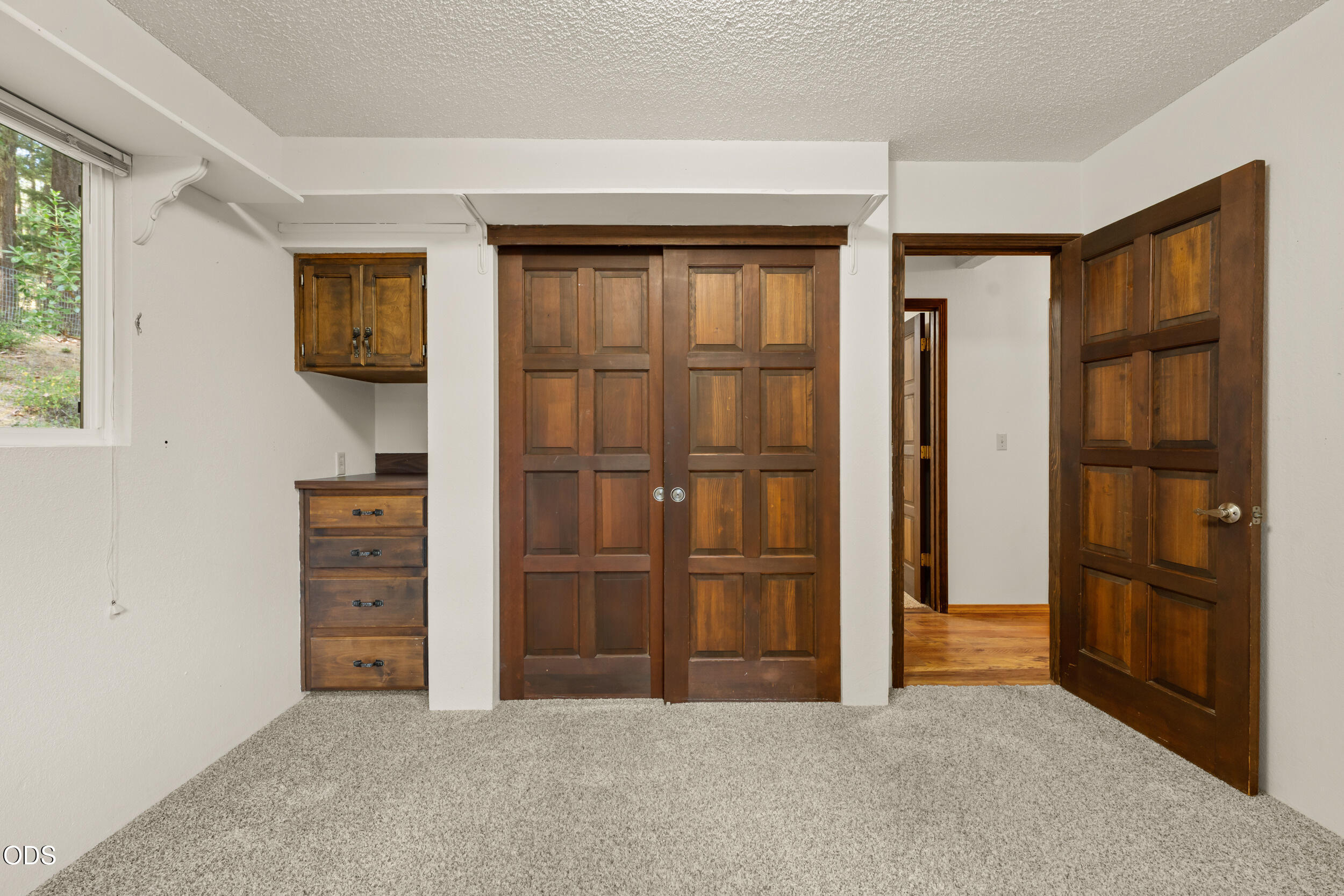 Carpeted bedroom with dark wood closet doors, built-in dresser, and window overlooking trees.