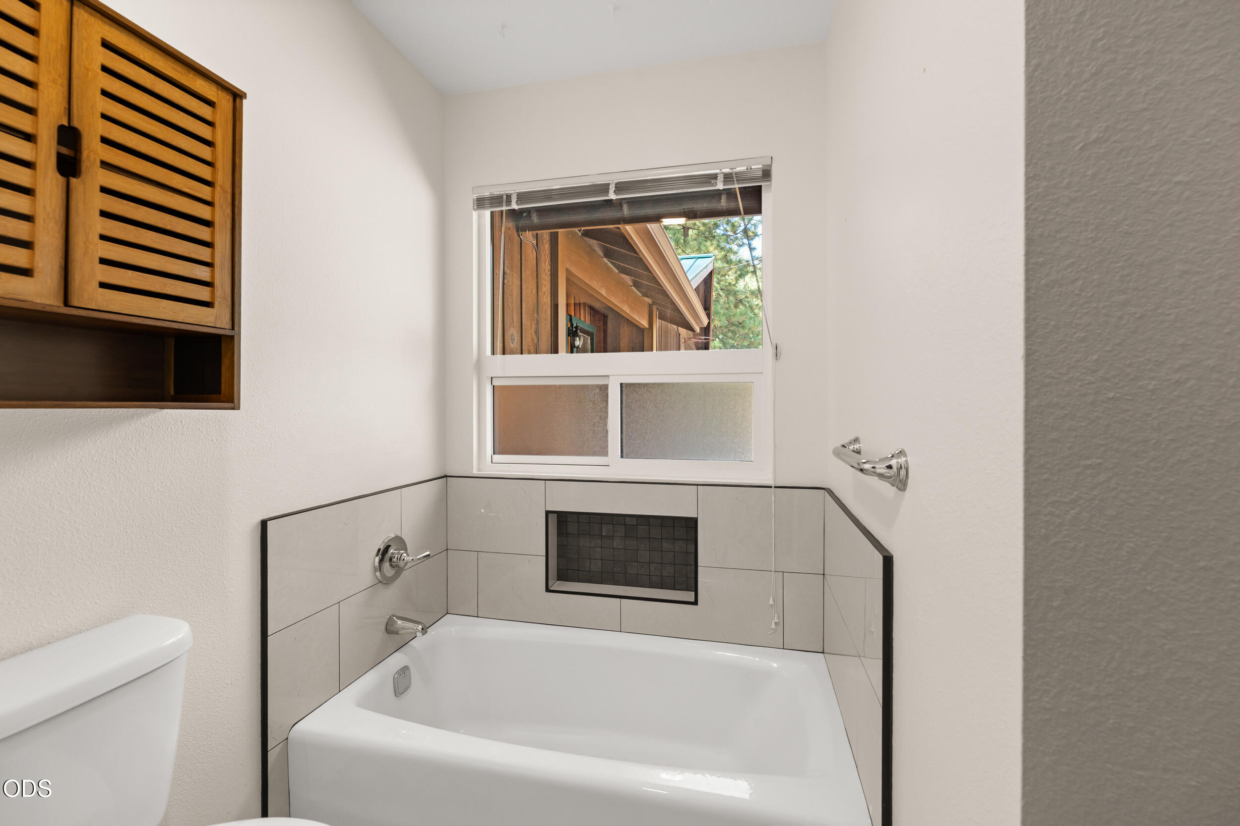 Bathroom with a white bathtub, tiled surround, wooden cabinet, and window.