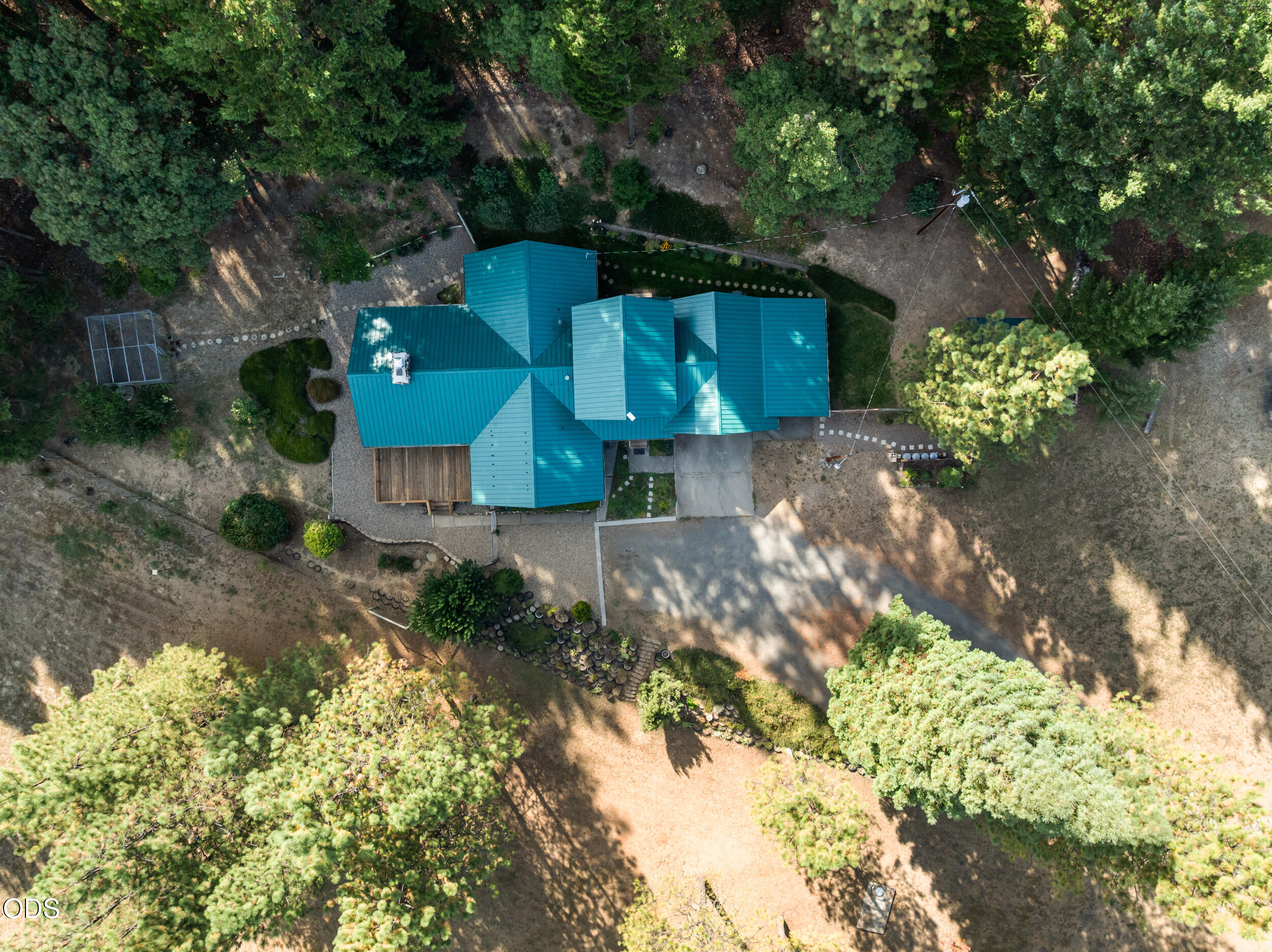 Aerial view of a teal-roofed house with a deck and driveway surrounded by mature trees.