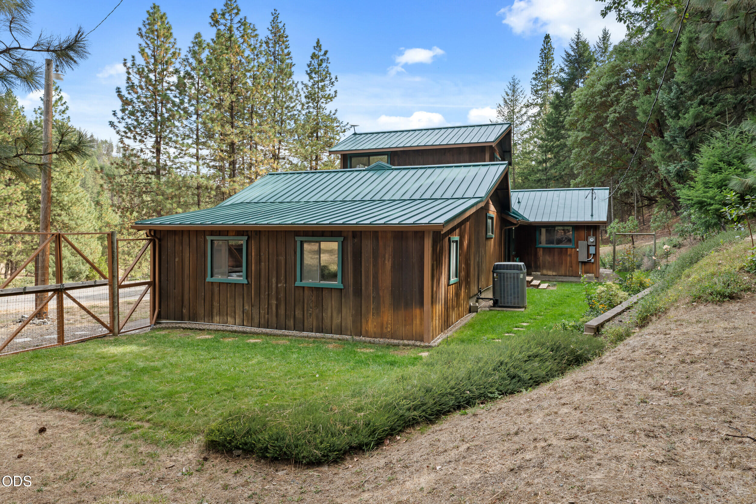 Wood-sided house with a green metal roof among pine trees, viewed beside a fenced gate.