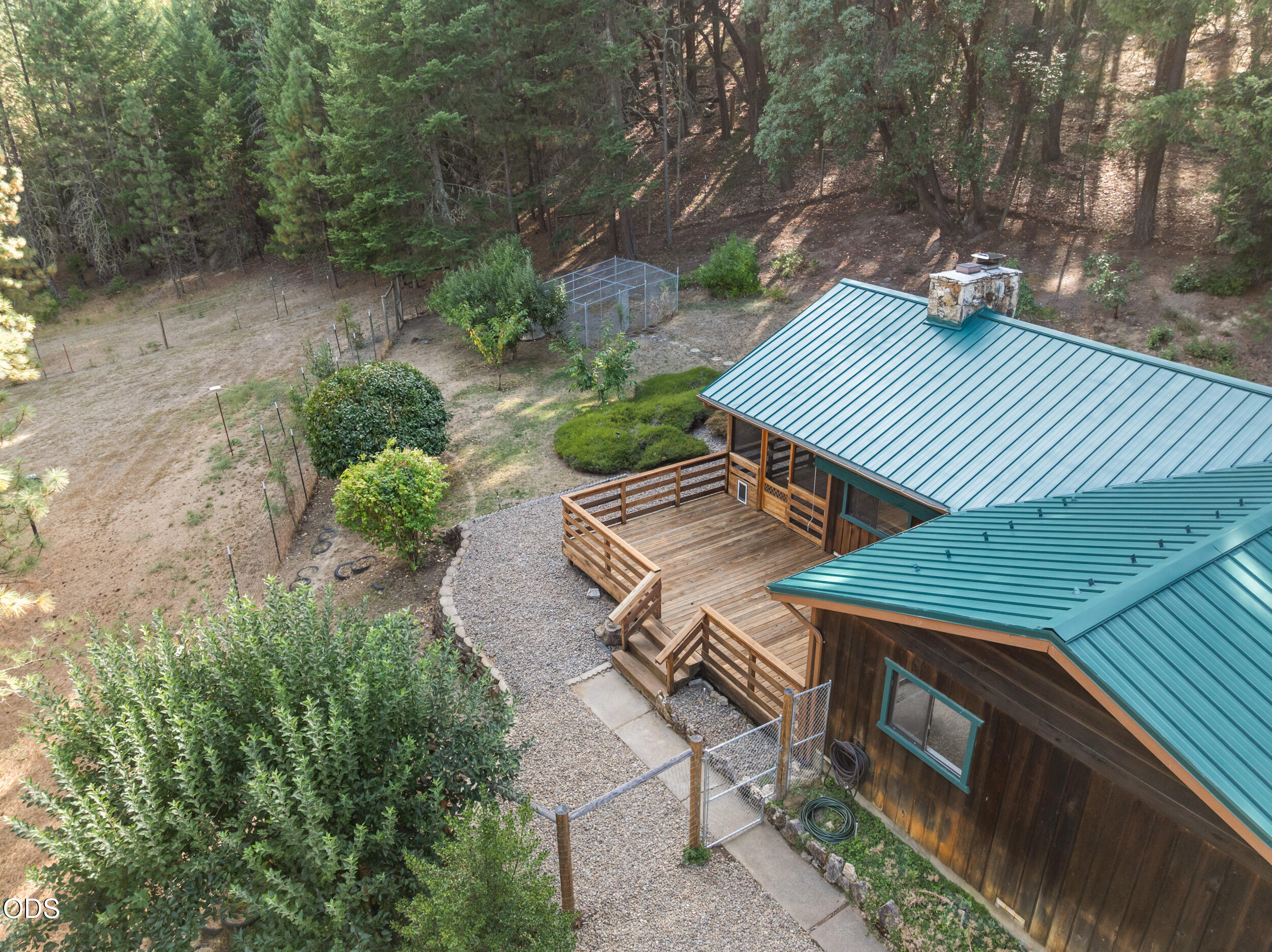 Aerial view of a wood-sided home with teal metal roofs, a broad deck, gravel paths, and wooded surroundings.