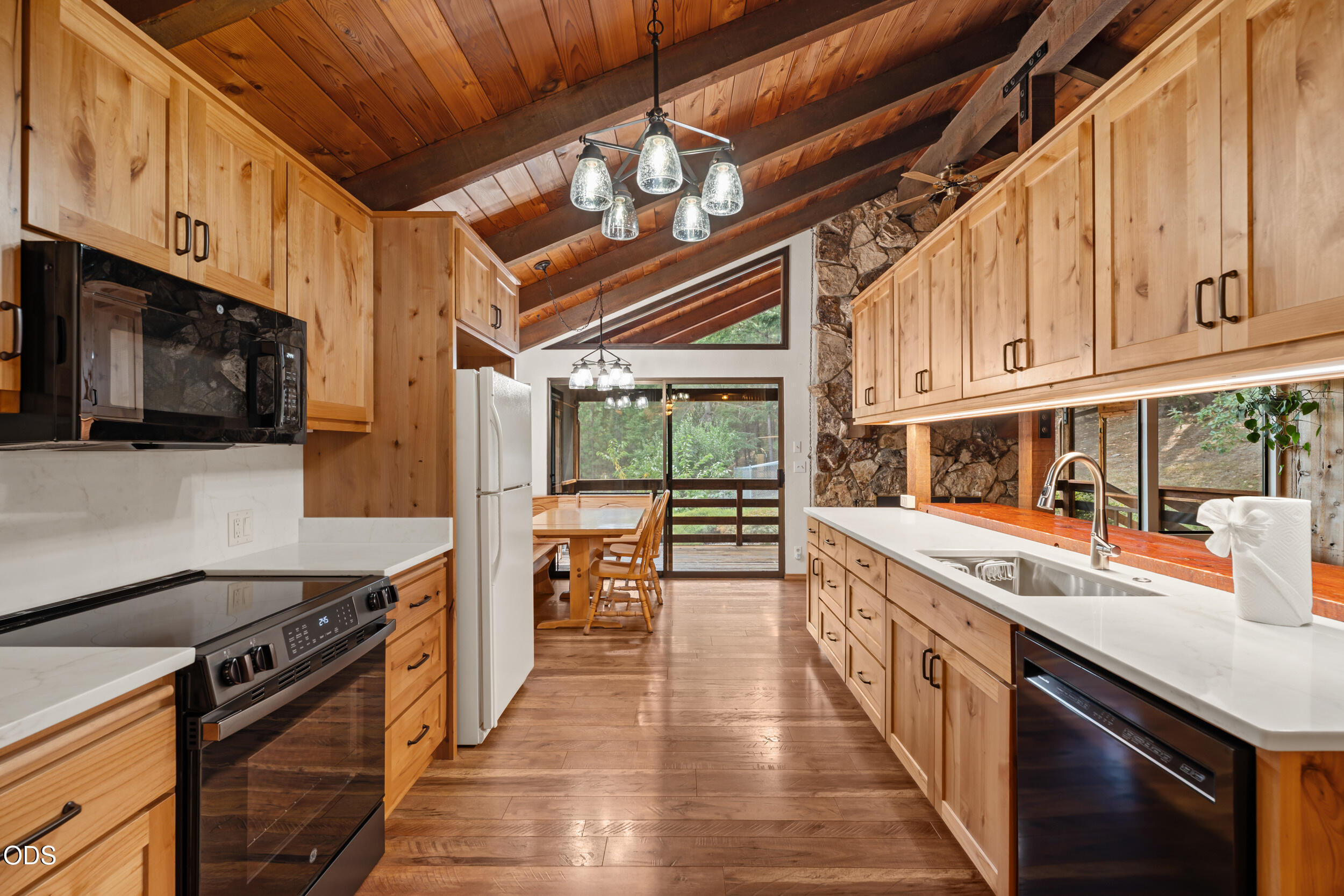 Kitchen with light wood cabinetry, vaulted beam ceiling, stone wall, and windows overlooking trees.