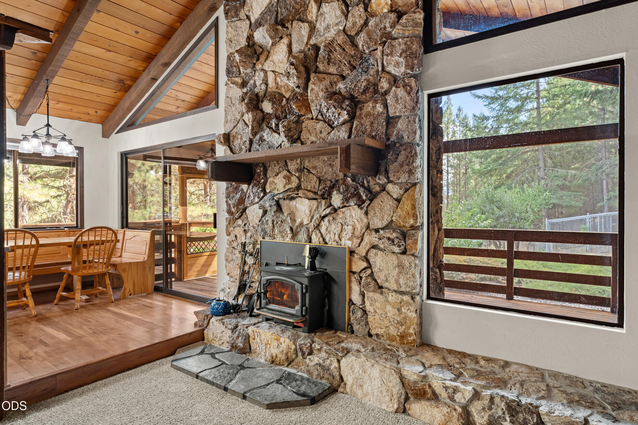 Living and dining area with a tall stone fireplace, wood-burning stove, vaulted ceiling, and forest views.