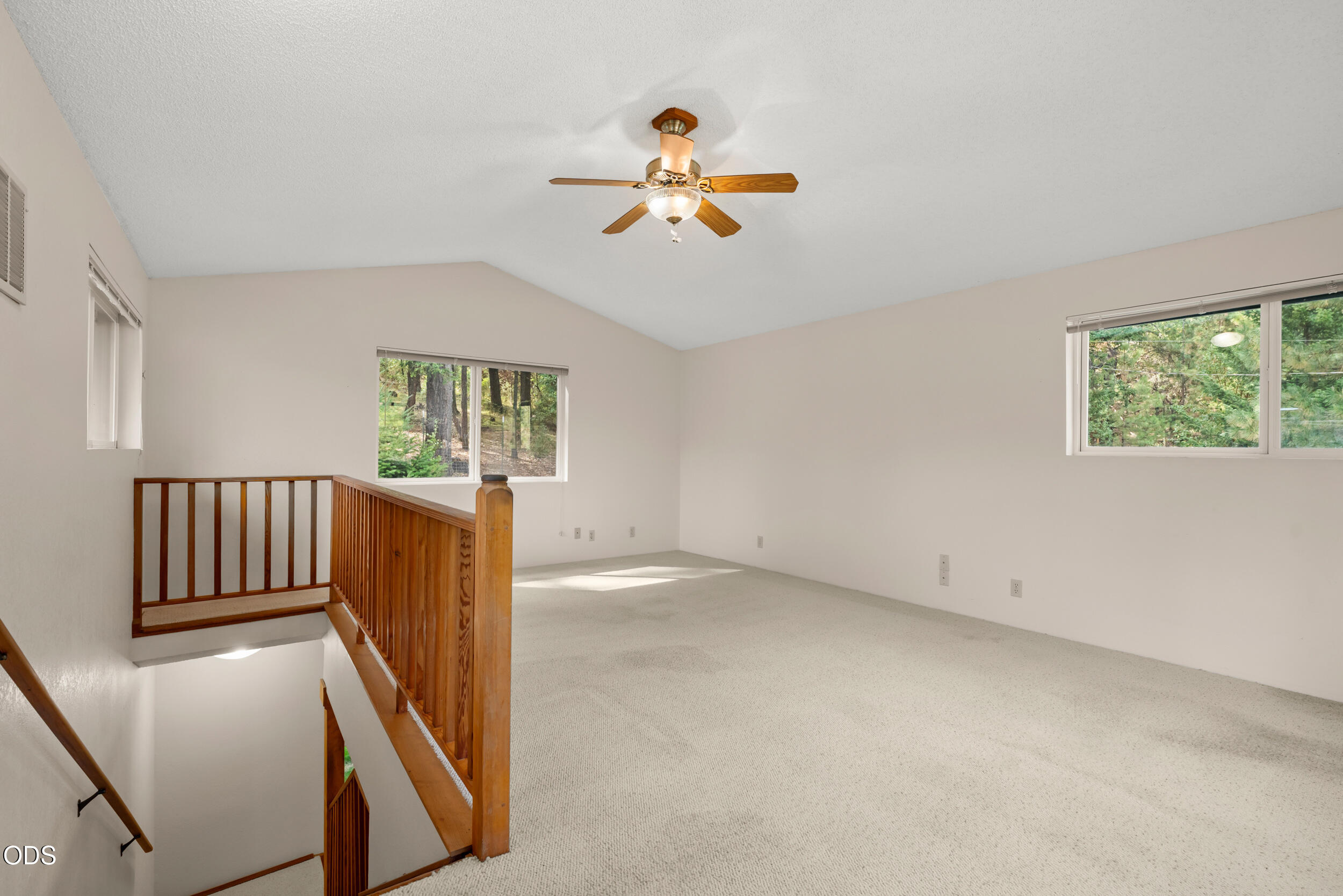 Vacant upstairs room with a ceiling fan, wooded views, and a wooden railing overlooking the stairs.