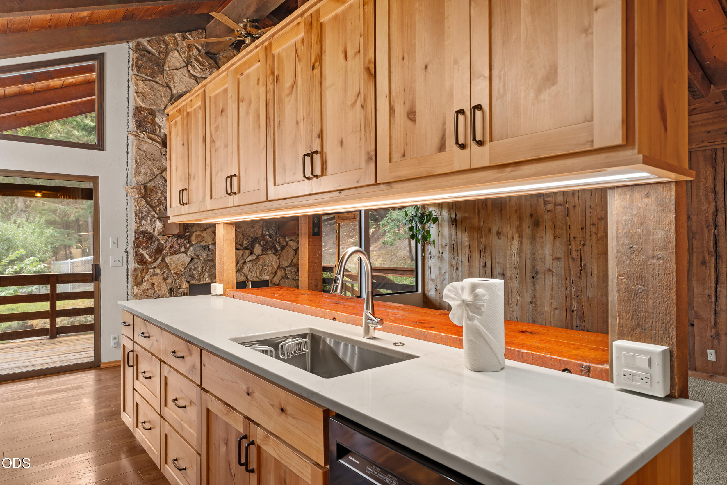 Kitchen with light wood cabinets, a white countertop, stainless-steel sink, and rustic stone and wood walls.