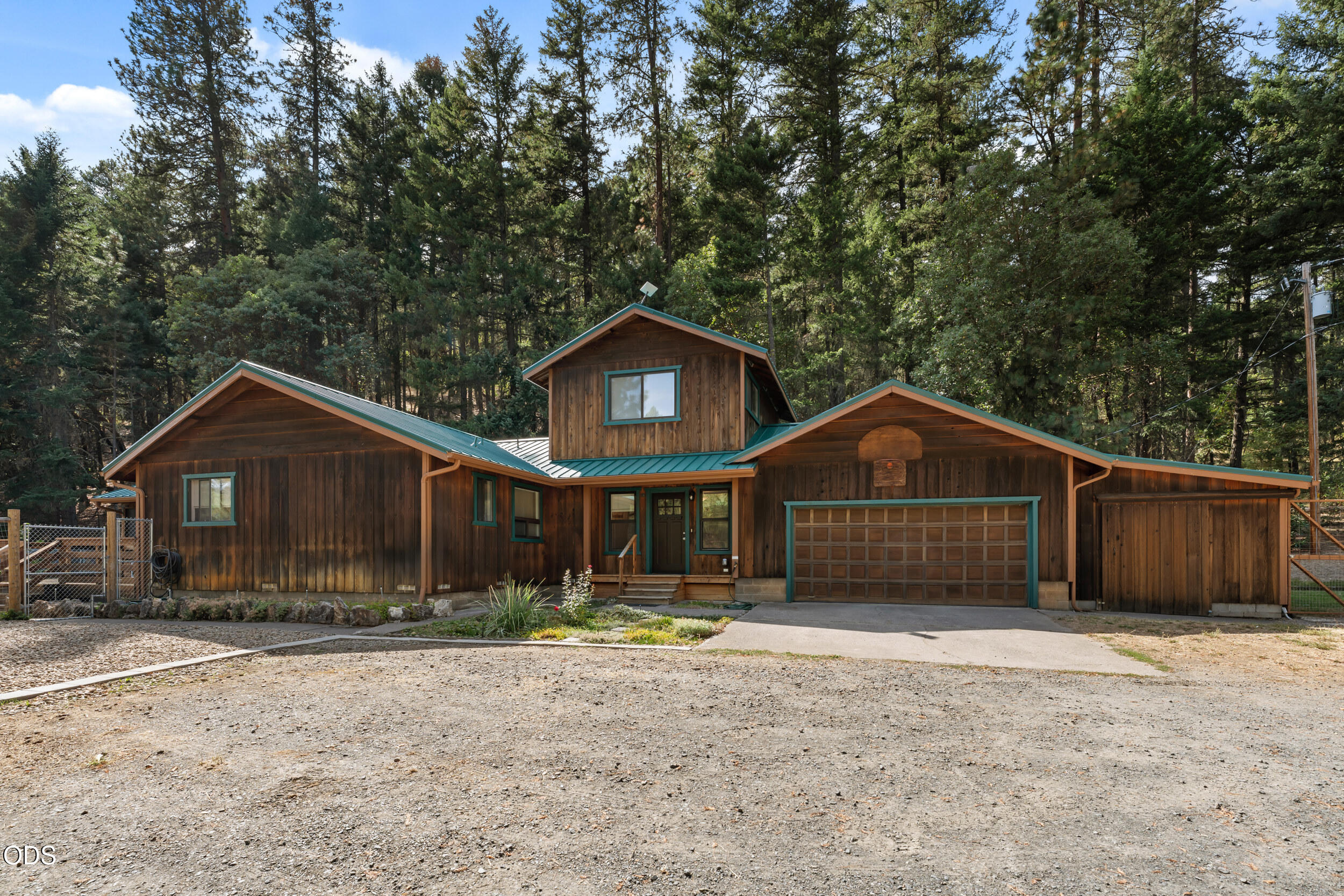 Wood-sided home with teal metal roofs, two-car garage, gravel driveway, and evergreen forest backdrop.