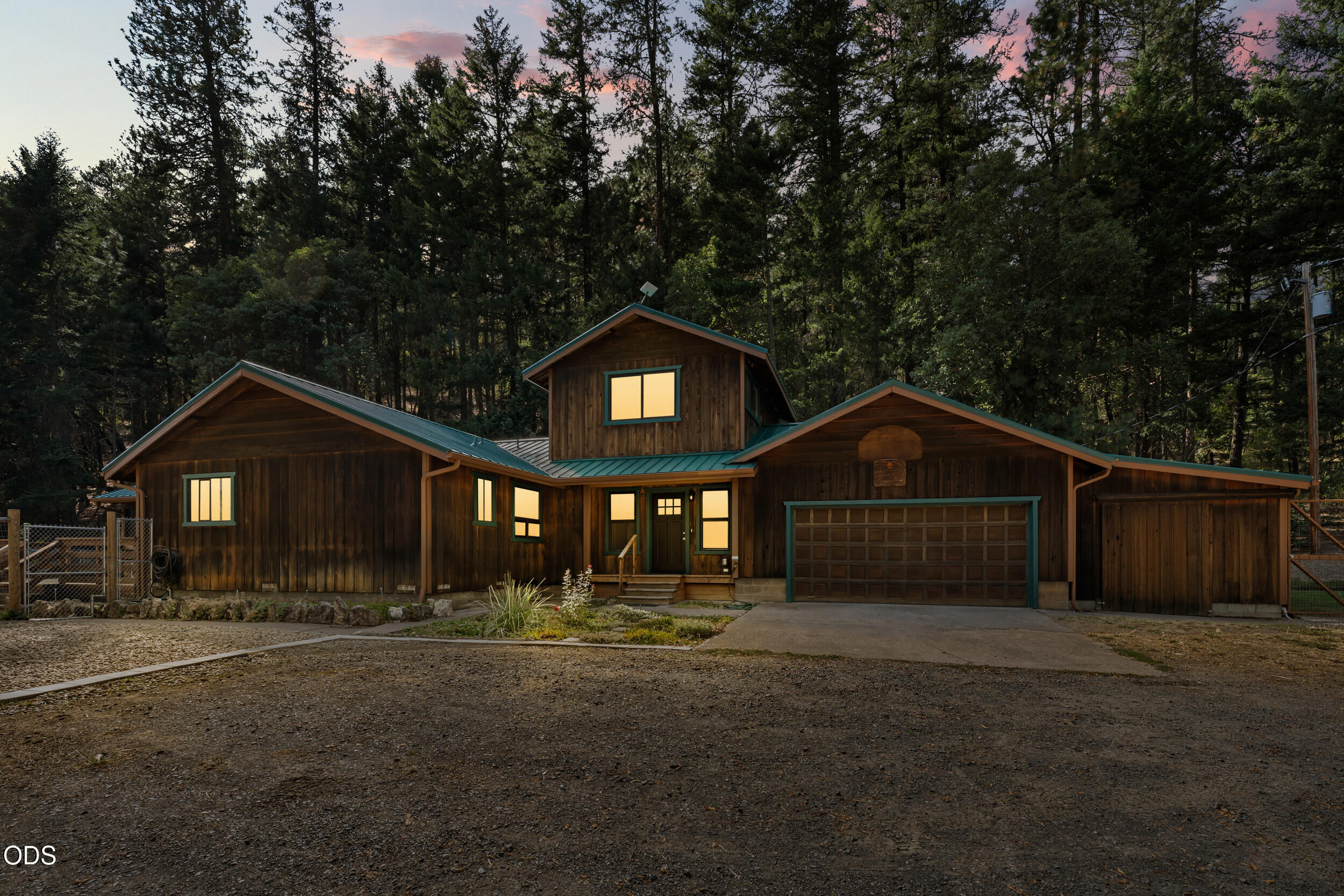 Wood-sided home with an attached garage and glowing windows, surrounded by evergreen trees at dusk.