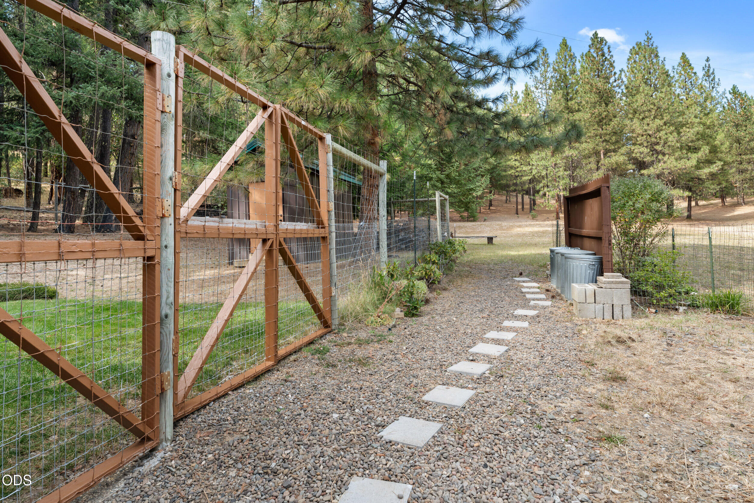 Gravel path with stepping stones leading through an open wire gate into a fenced, pine-shaded yard.