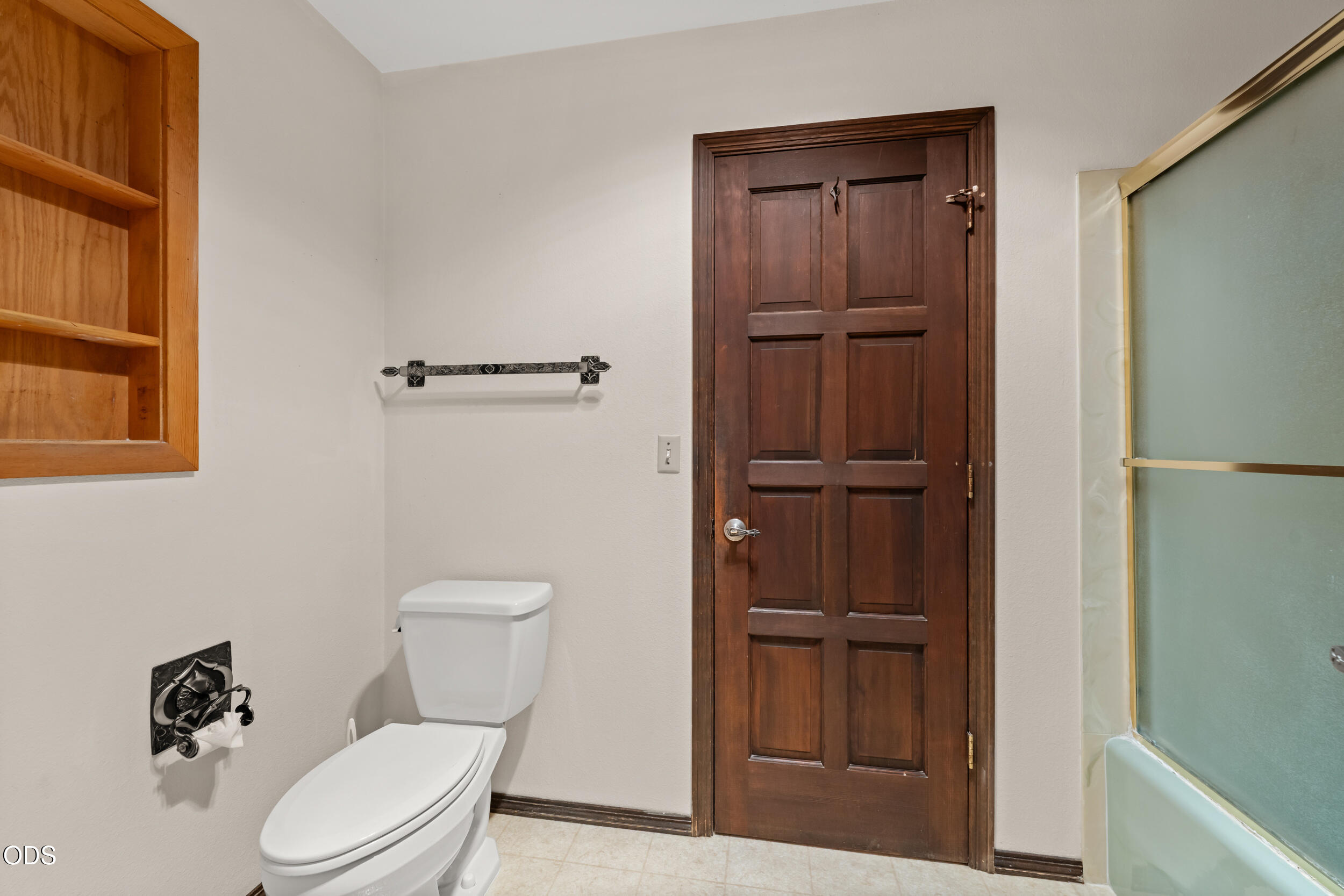 Bathroom with a dark wood paneled door, built-in shelves, and frosted-glass tub enclosure.