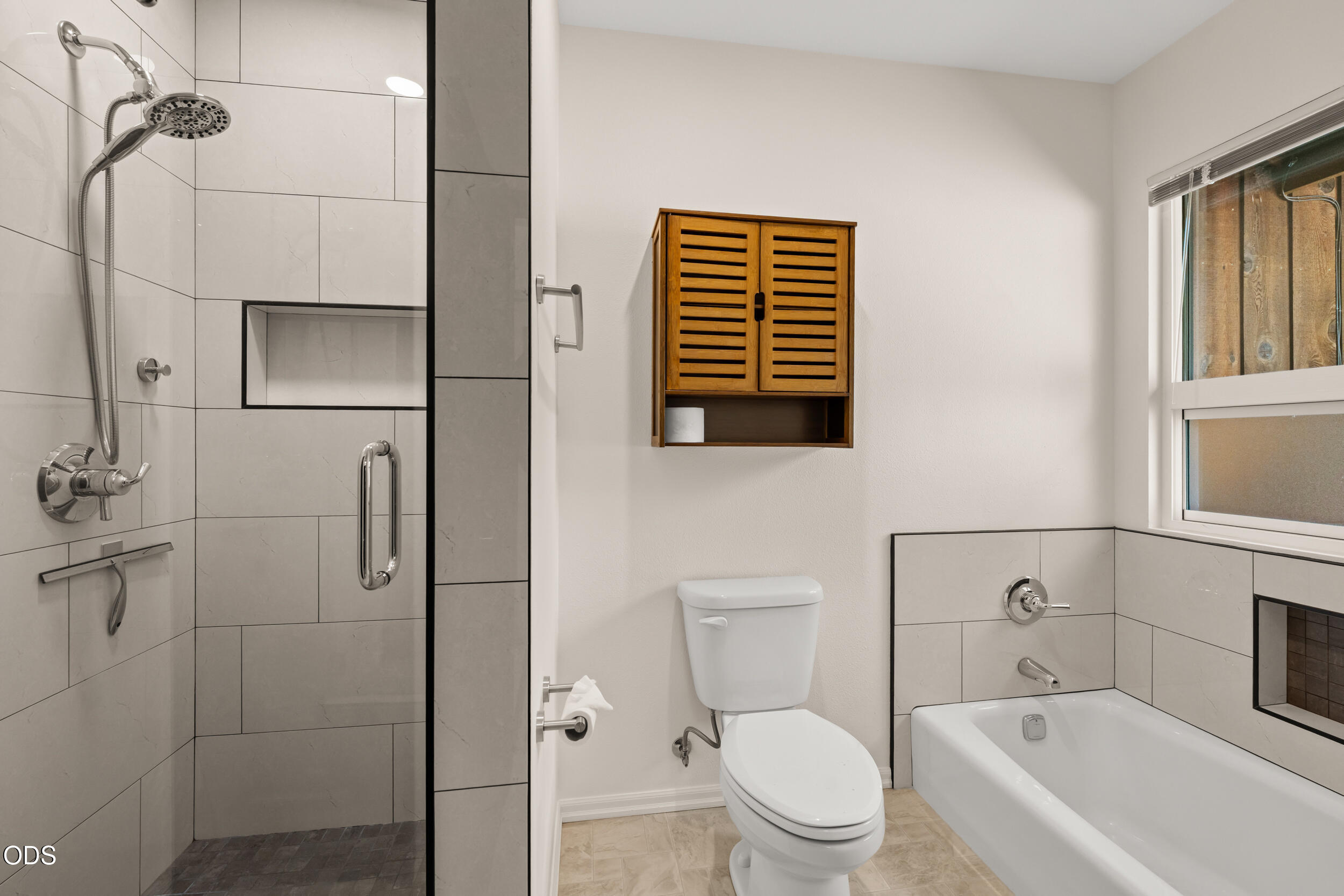 Bathroom with a glass-enclosed tiled shower, bathtub beneath a window, and wooden wall cabinet.