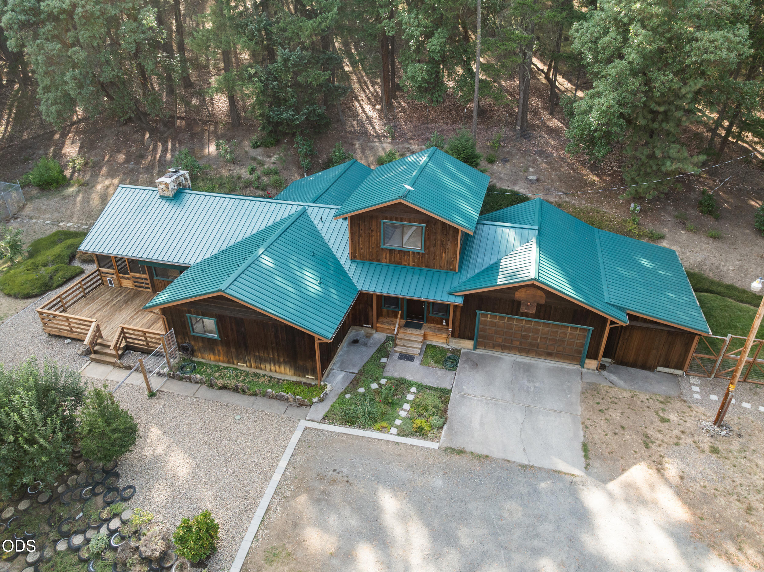 Aerial view of a wood-sided home with teal metal roofs, a front garage, and a deck beside a wooded lot.