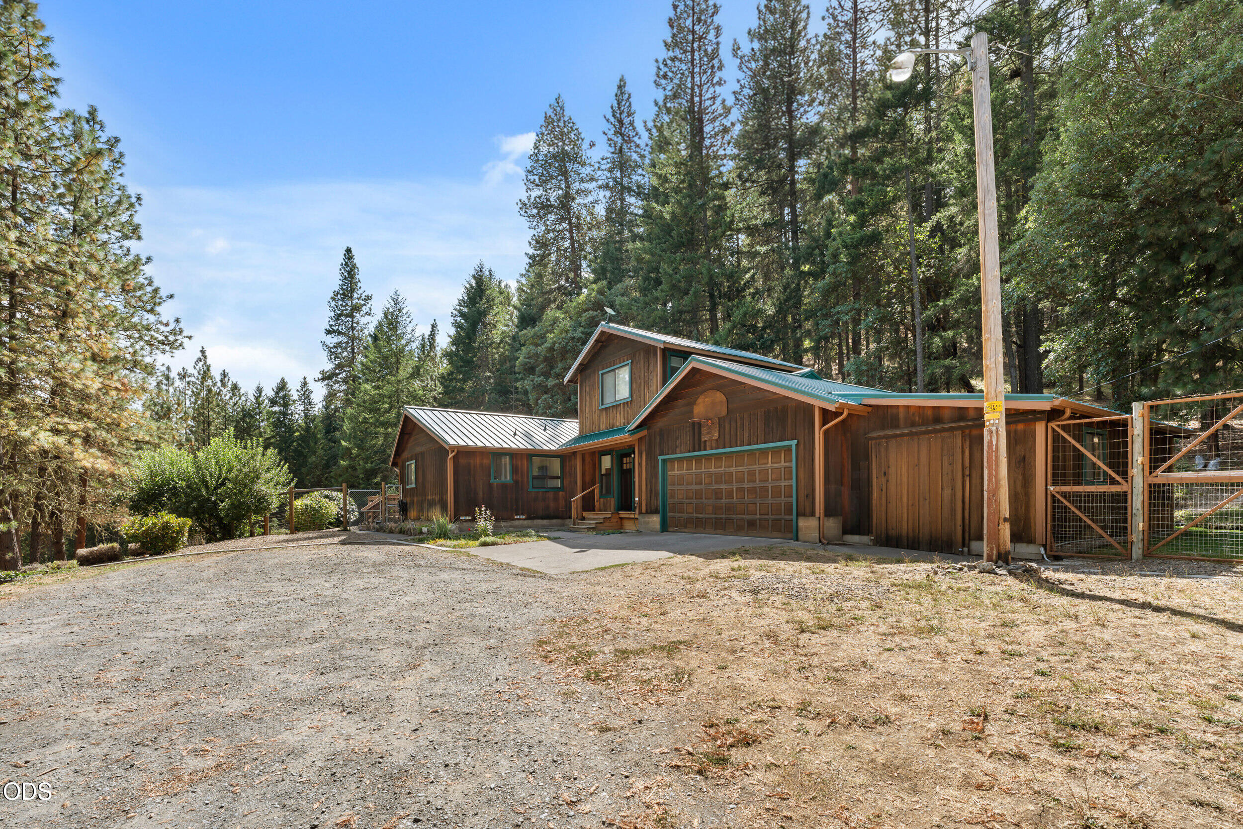 Wood-sided home with teal trim and a two-car garage among tall evergreen trees.