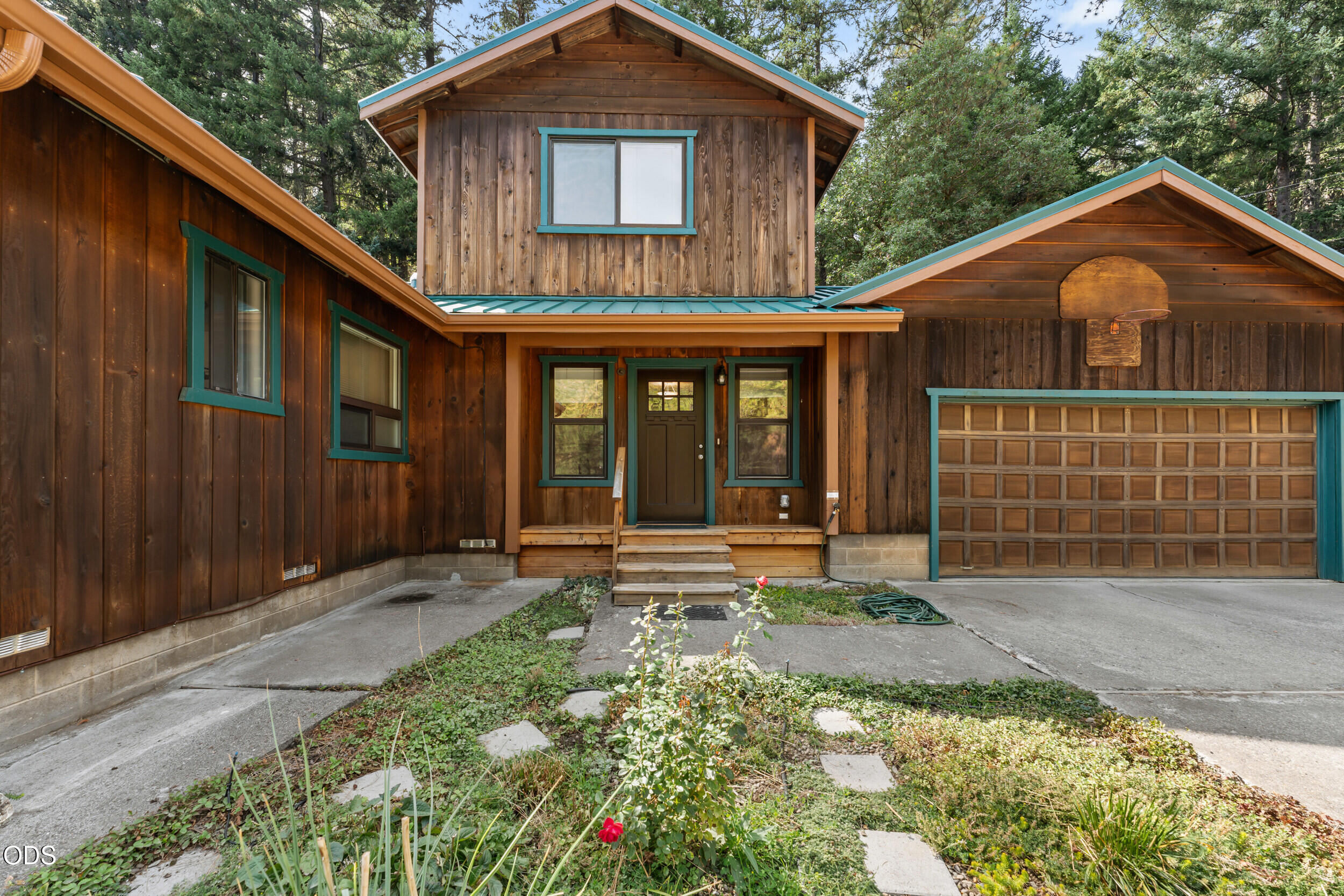 Wood-sided home with teal trim, covered entry, and attached double garage surrounded by trees.
