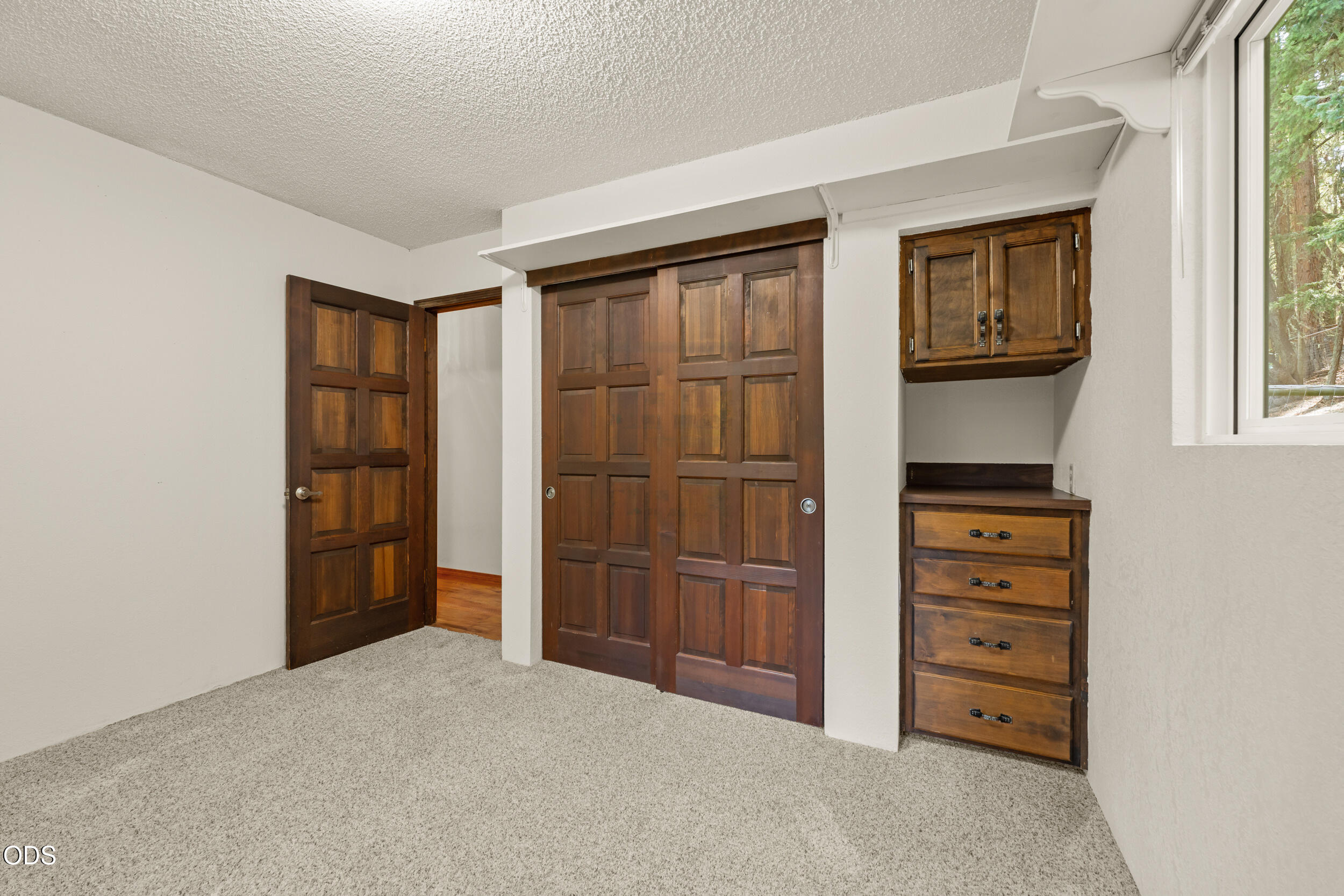 Empty bedroom with dark wood closet doors and built-in dresser beside a window.