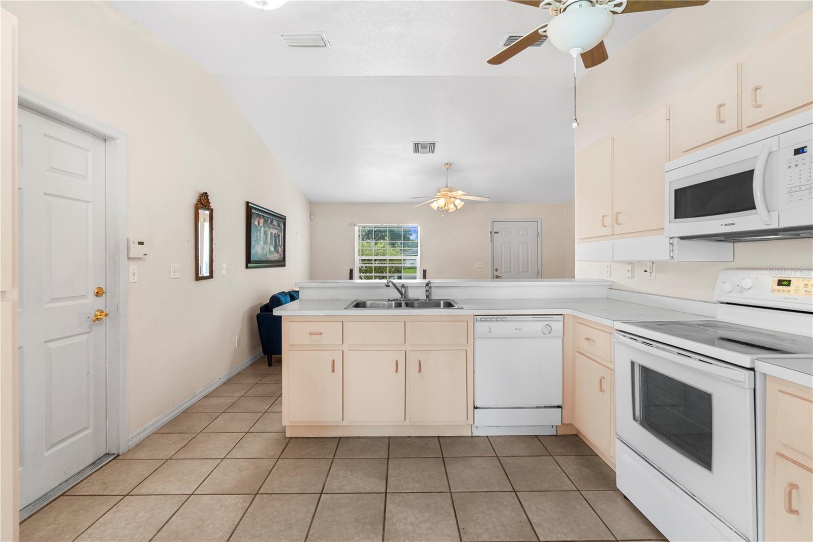 Kitchen with a peninsula sink, pale cabinets, white appliances, and tan tile flooring.