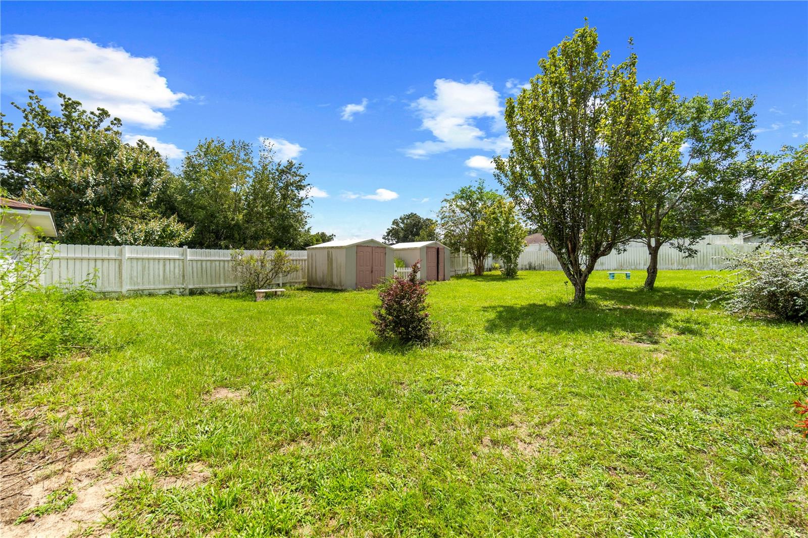 Backyard with a broad lawn, mature trees, white privacy fence, and two storage sheds.