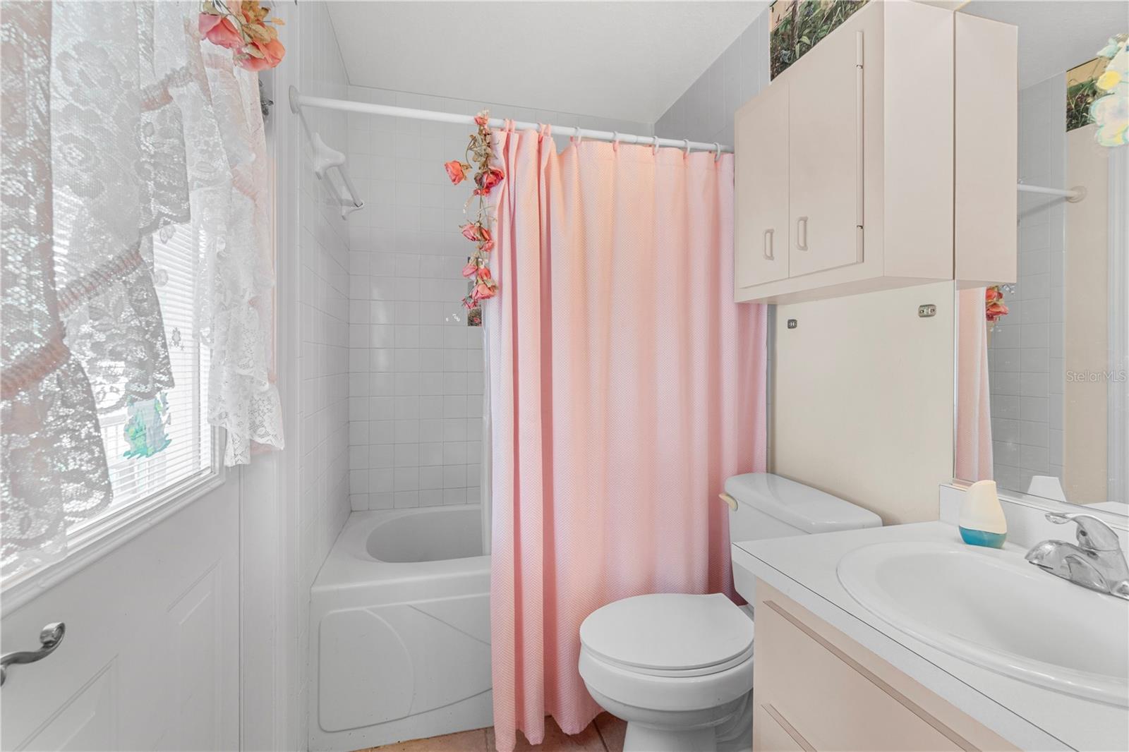 Bathroom with a pink shower curtain, bathtub, toilet, and white vanity beside a curtained window.
