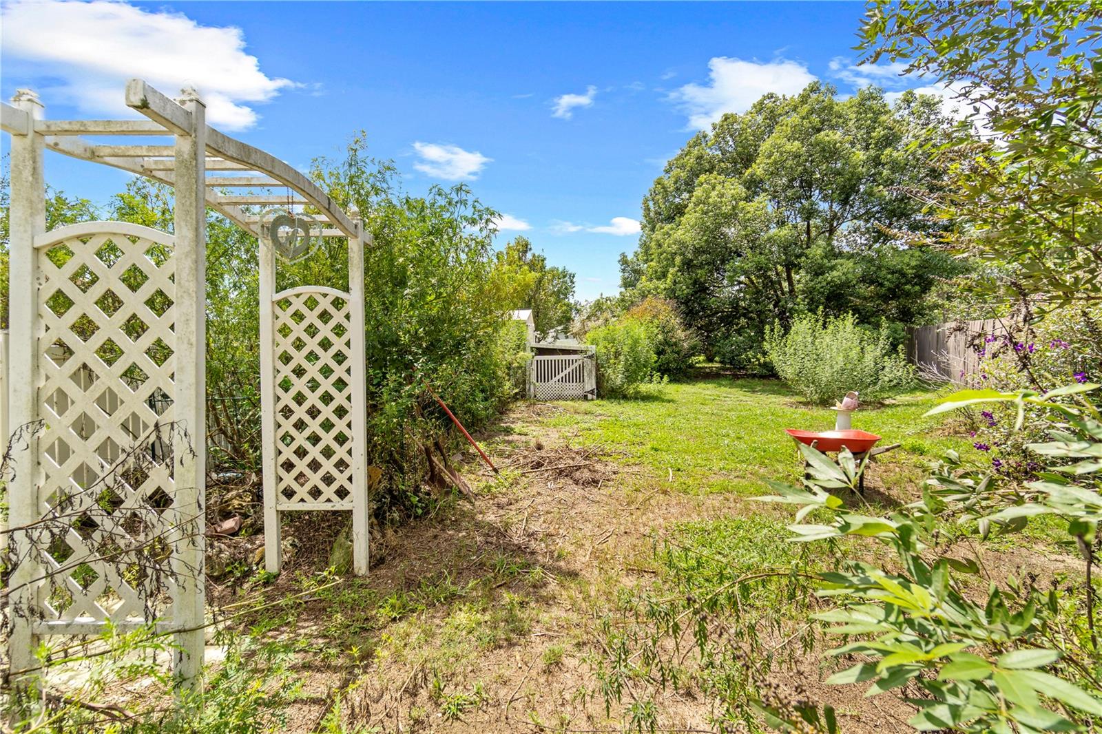 Backyard garden with a white lattice arbor, grassy clearing, mature trees, and fenced perimeter.