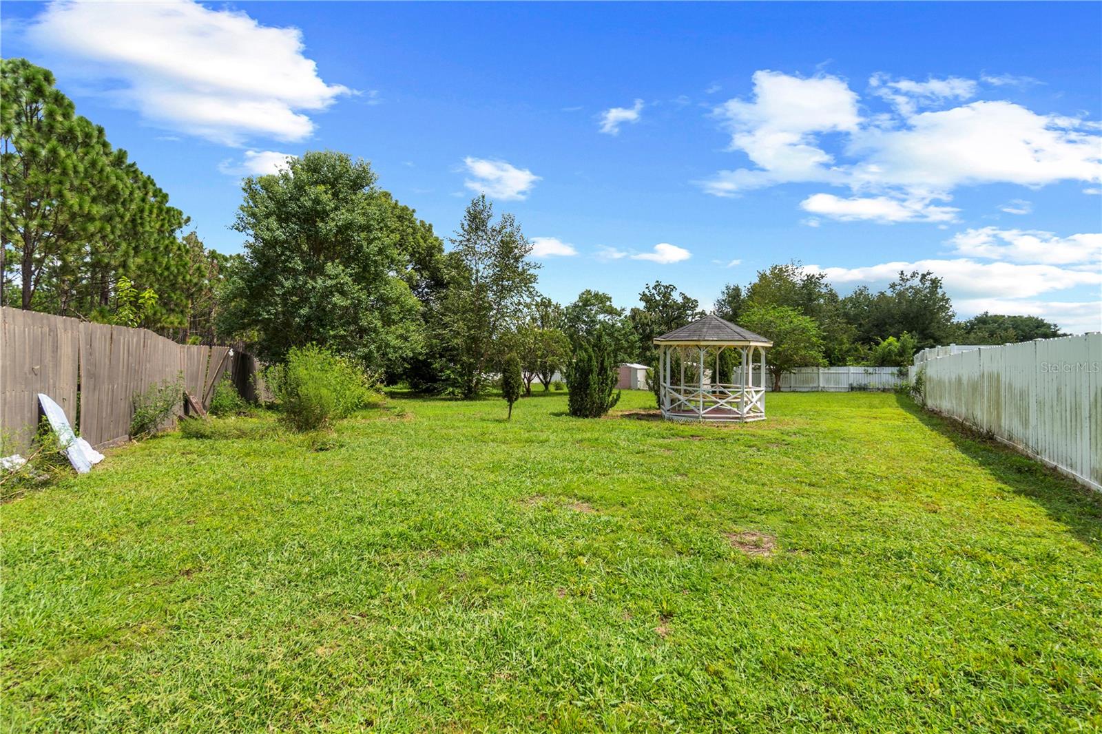 Fenced grassy backyard with mature trees and a small white gazebo.