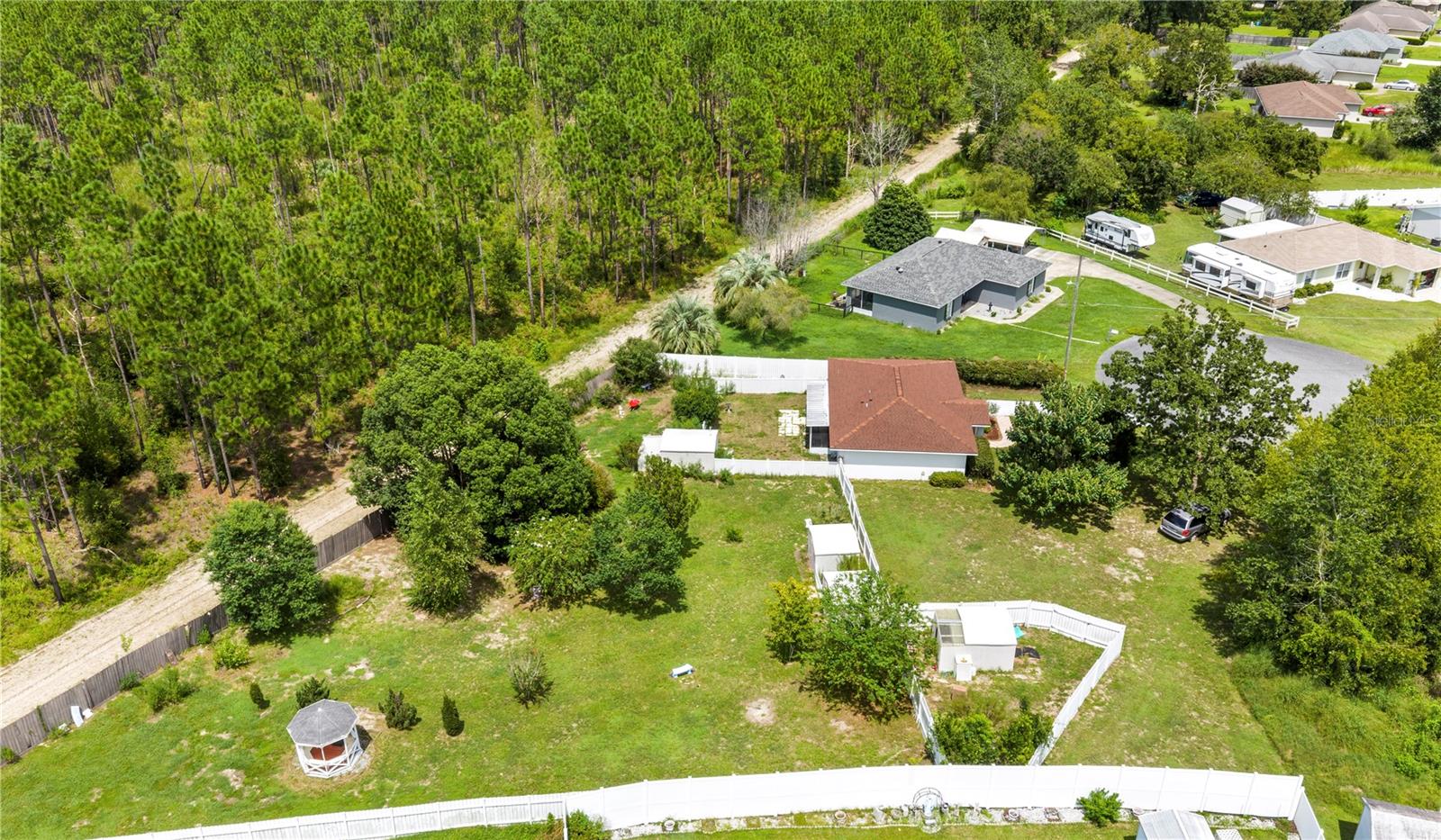Aerial view of a home with a reddish-brown roof, fenced yard, gazebo, and bordering pine woods.