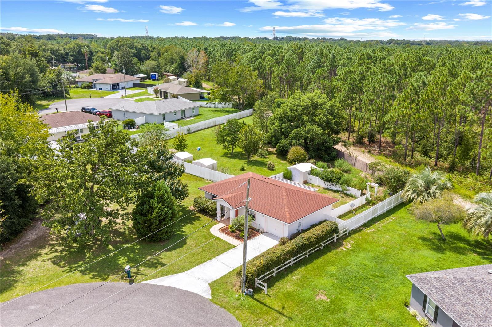 Single-story home with a red tile roof and fenced yard beside a pine forest.