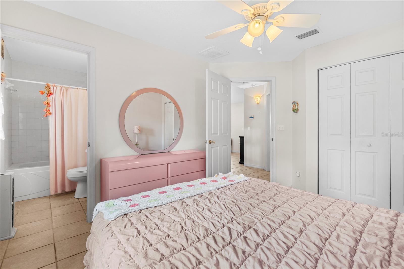 Bedroom with a pink dresser and round mirror, ceiling fan, and adjoining bathroom with a tub and shower.