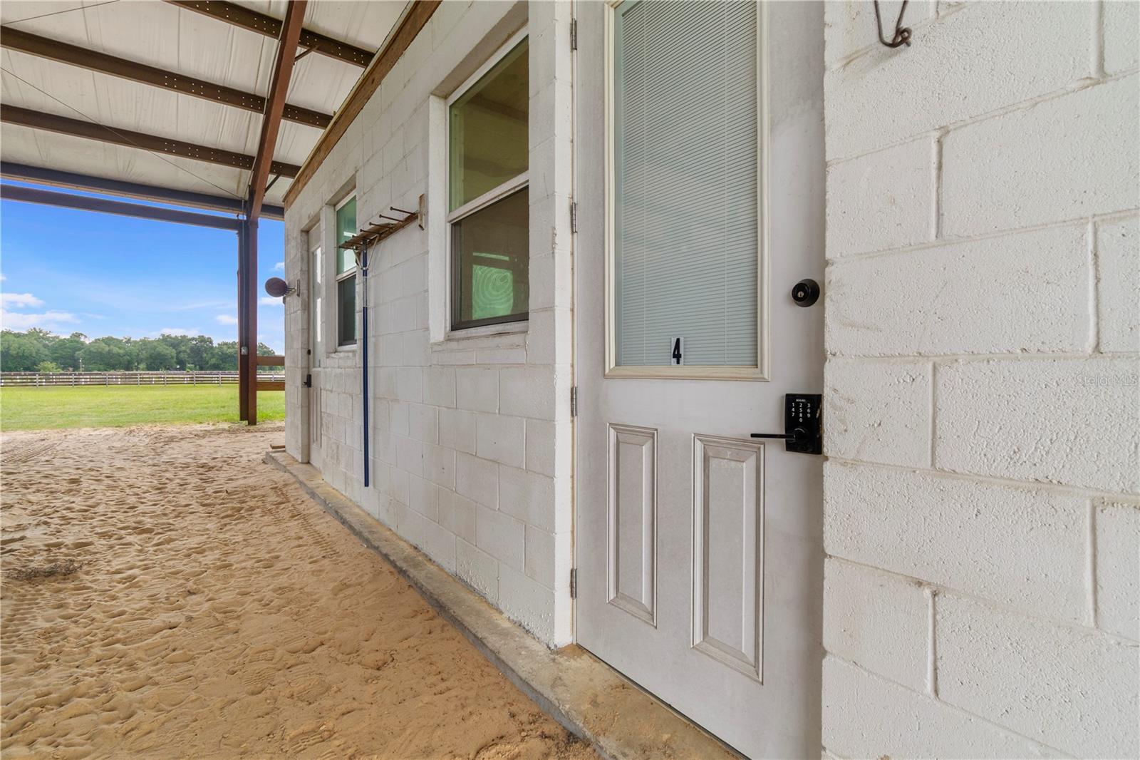 White block building entrance with a numbered door beside a covered sandy walkway and fenced field.