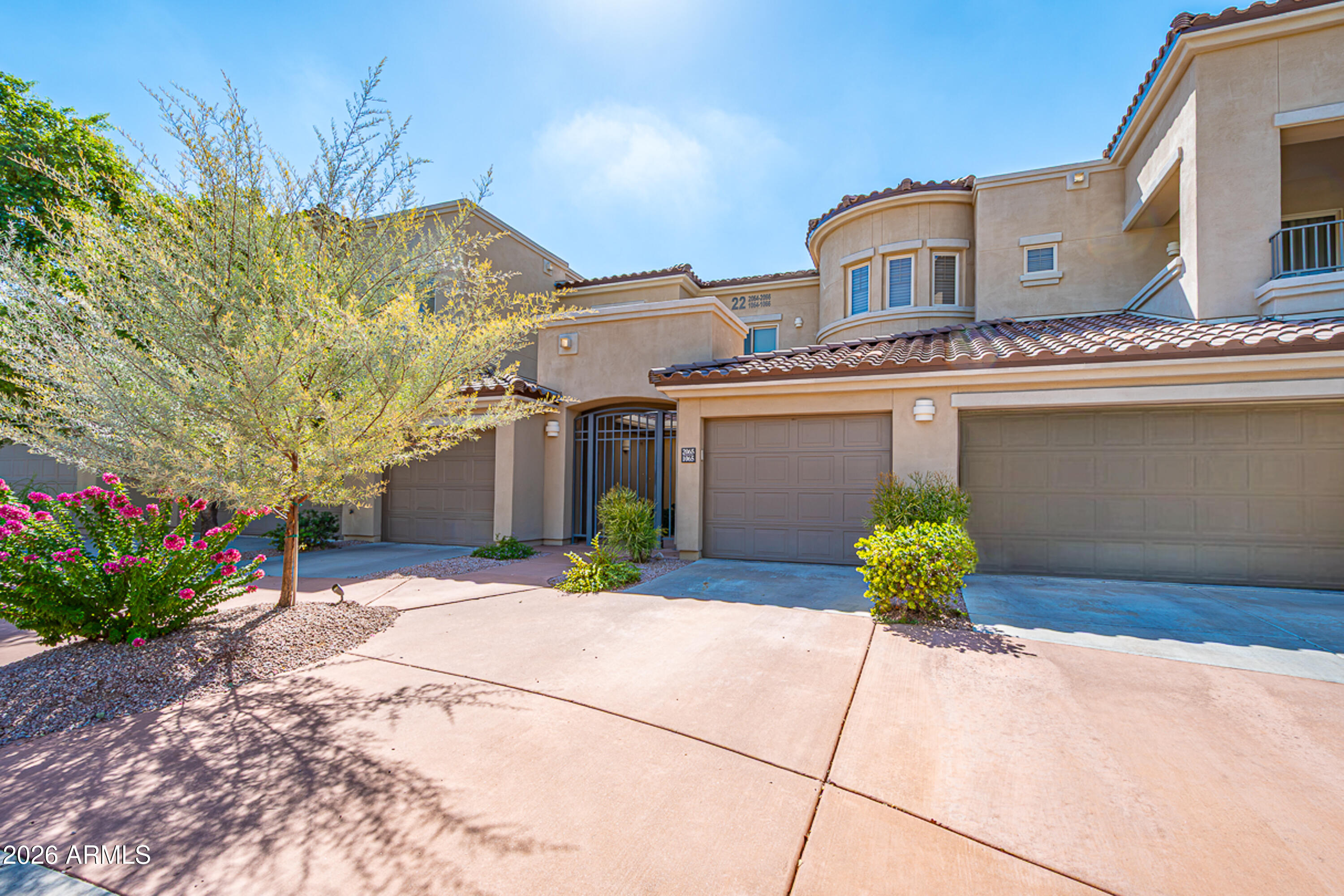 Two-story beige home with tile roofs, multiple garage doors, an arched entry, and desert landscaping.