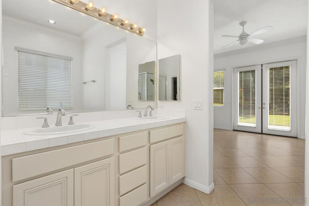 Bathroom with a long double-sink vanity and large mirror, opening to a tiled room with French doors.