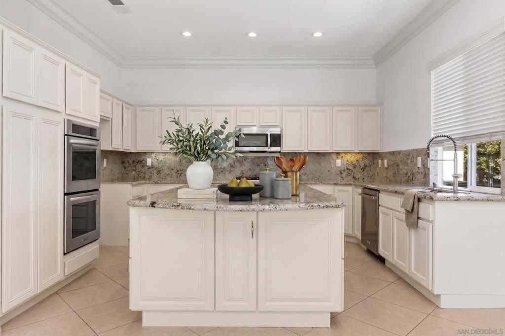 Bright kitchen with a large granite island, white cabinetry, stainless steel appliances, and a sink beneath a window.