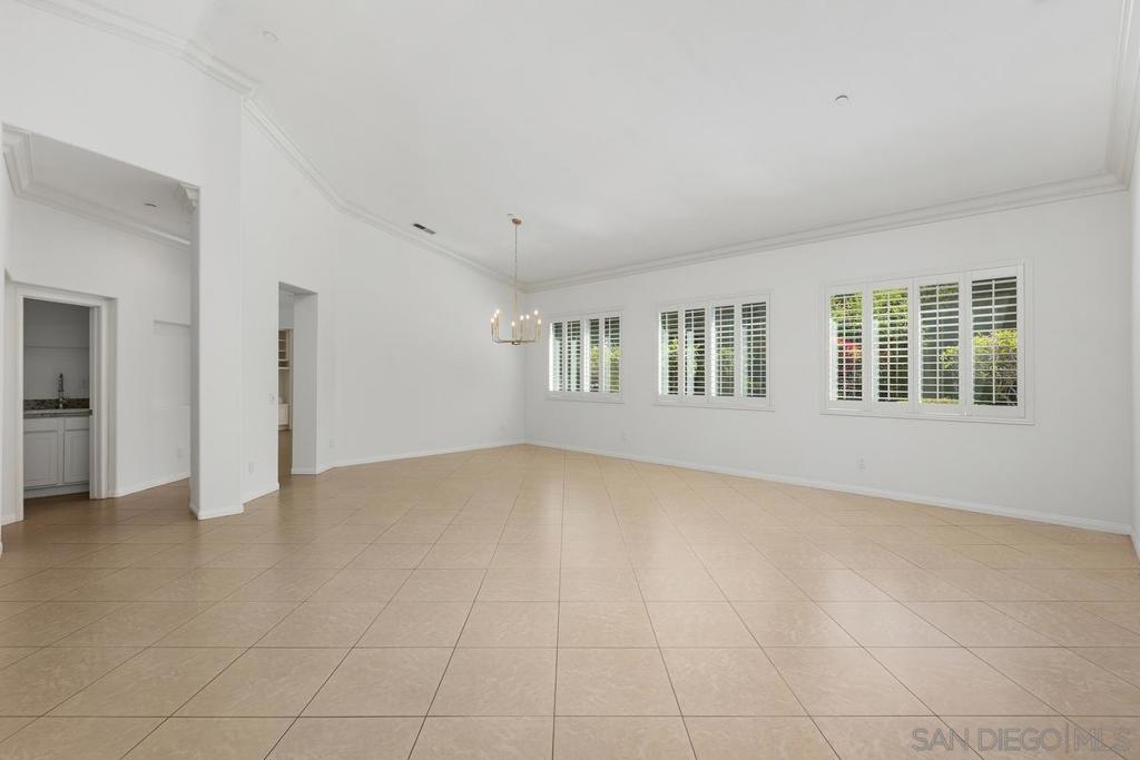 Empty spacious room with beige tile floors, a chandelier, and shuttered windows.