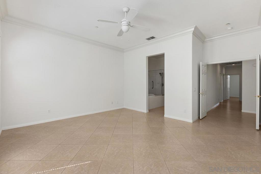 Empty bedroom with beige tile floors, a ceiling fan, and doorways to an en-suite bathroom and hallway.