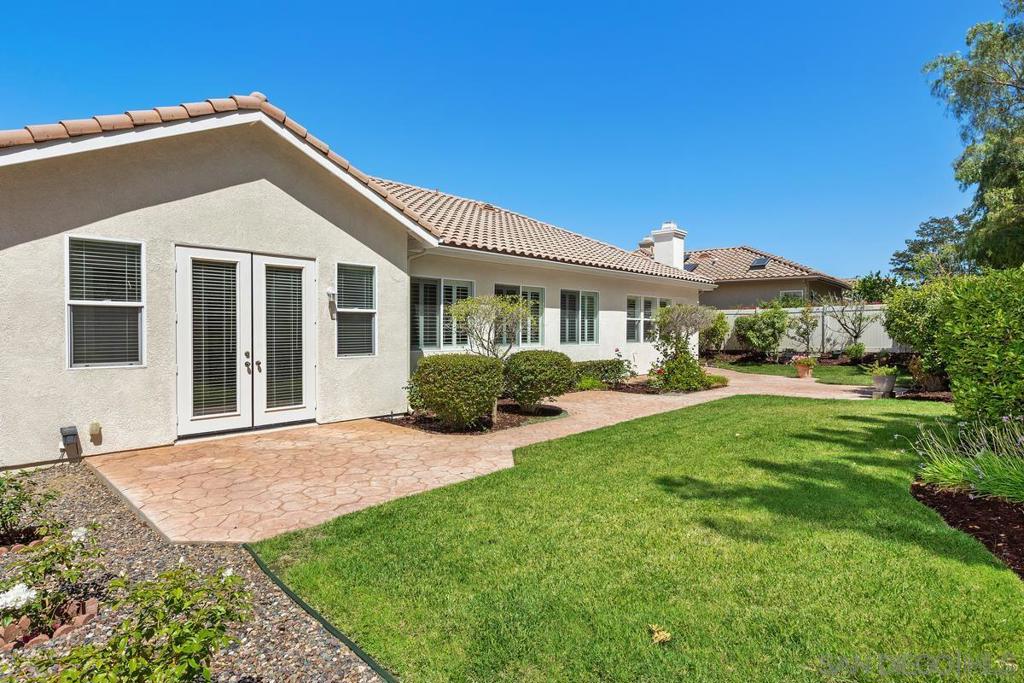 Rear of a stucco home with a tile roof, paved patio, trimmed shrubs, and a broad green lawn.