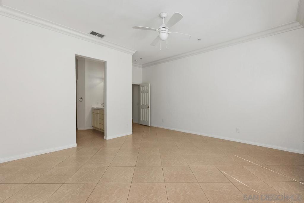 Empty bedroom with tan tile flooring, a ceiling fan, and an adjoining bathroom vanity.