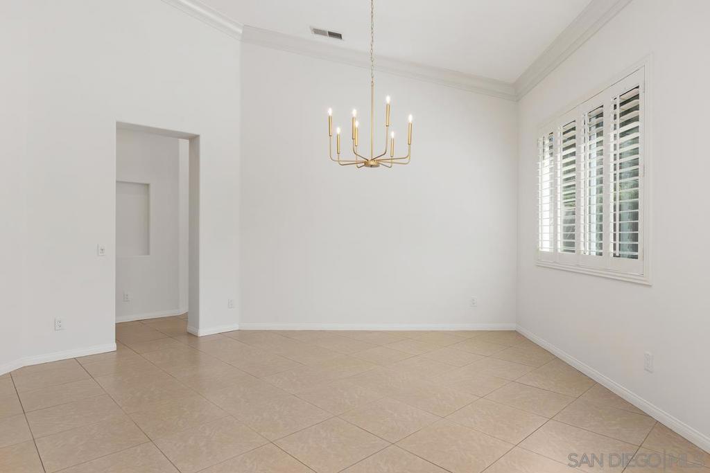 Empty dining room with beige tile floors, plantation shutters, and a brass chandelier.
