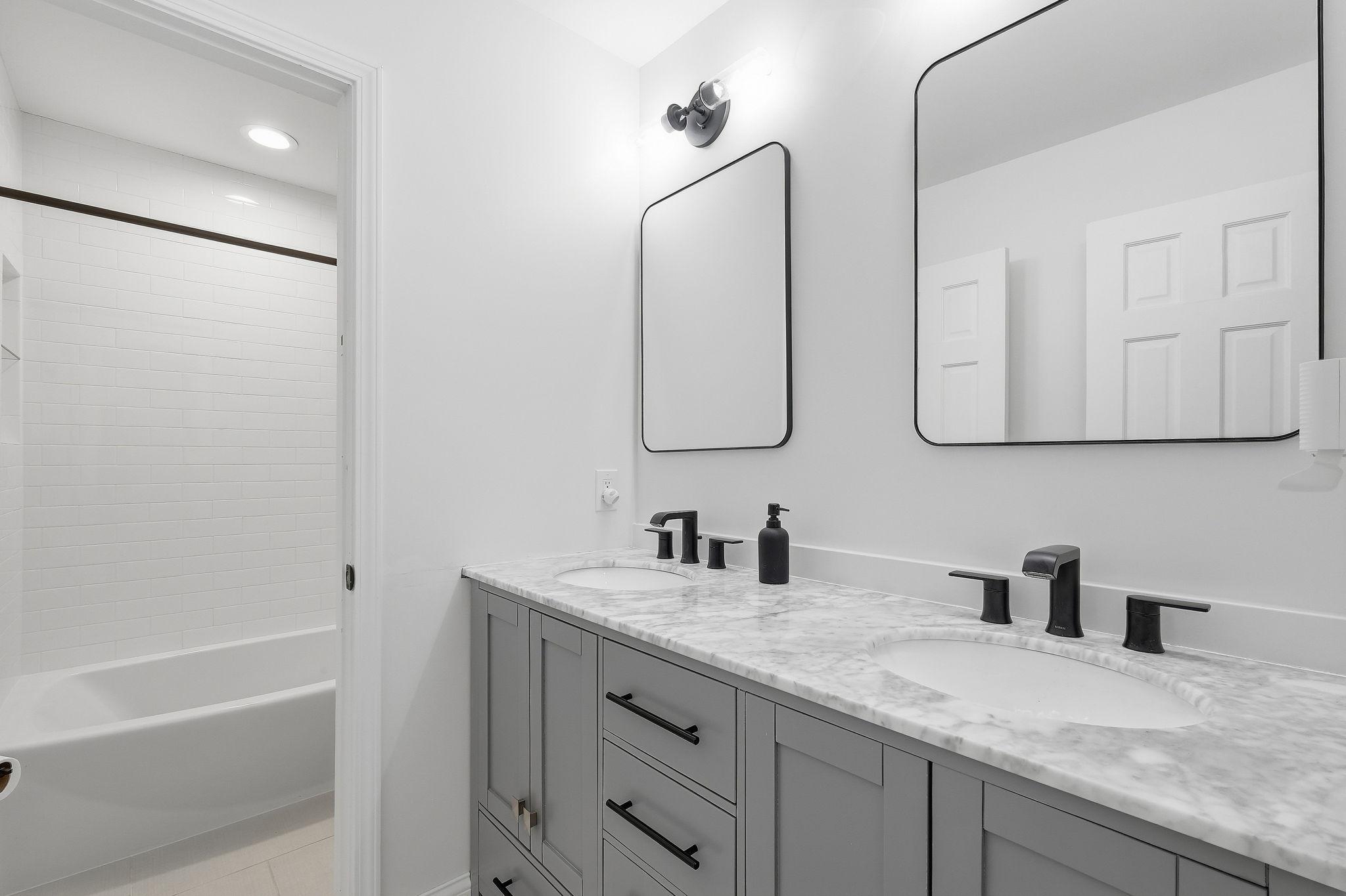 Bathroom with a double vanity, marble countertop, black faucets, and a subway-tile bathtub surround.