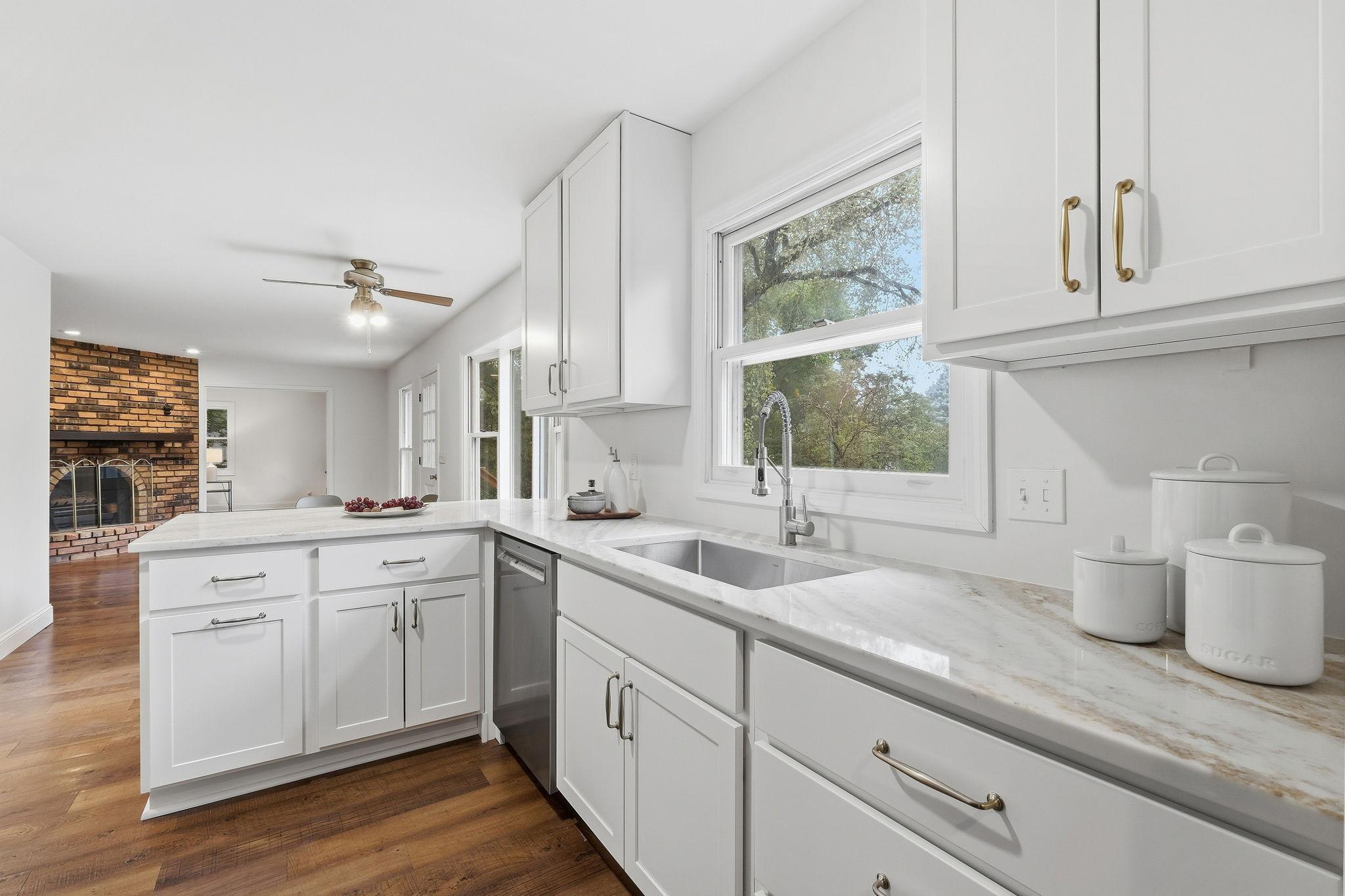 Bright white kitchen with marble-look counters, a large window, and an adjoining brick fireplace.