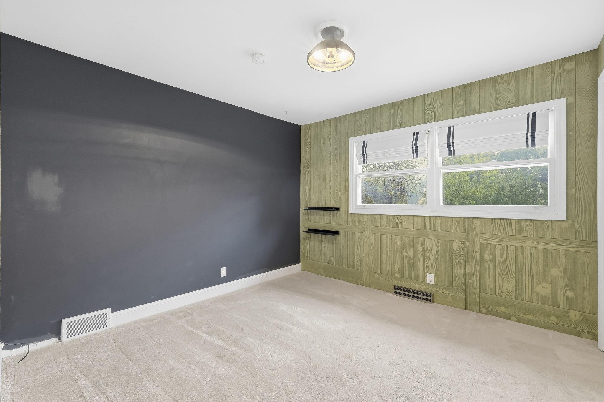 Empty carpeted room with a dark gray accent wall, green paneling, and a wide window with striped shades.