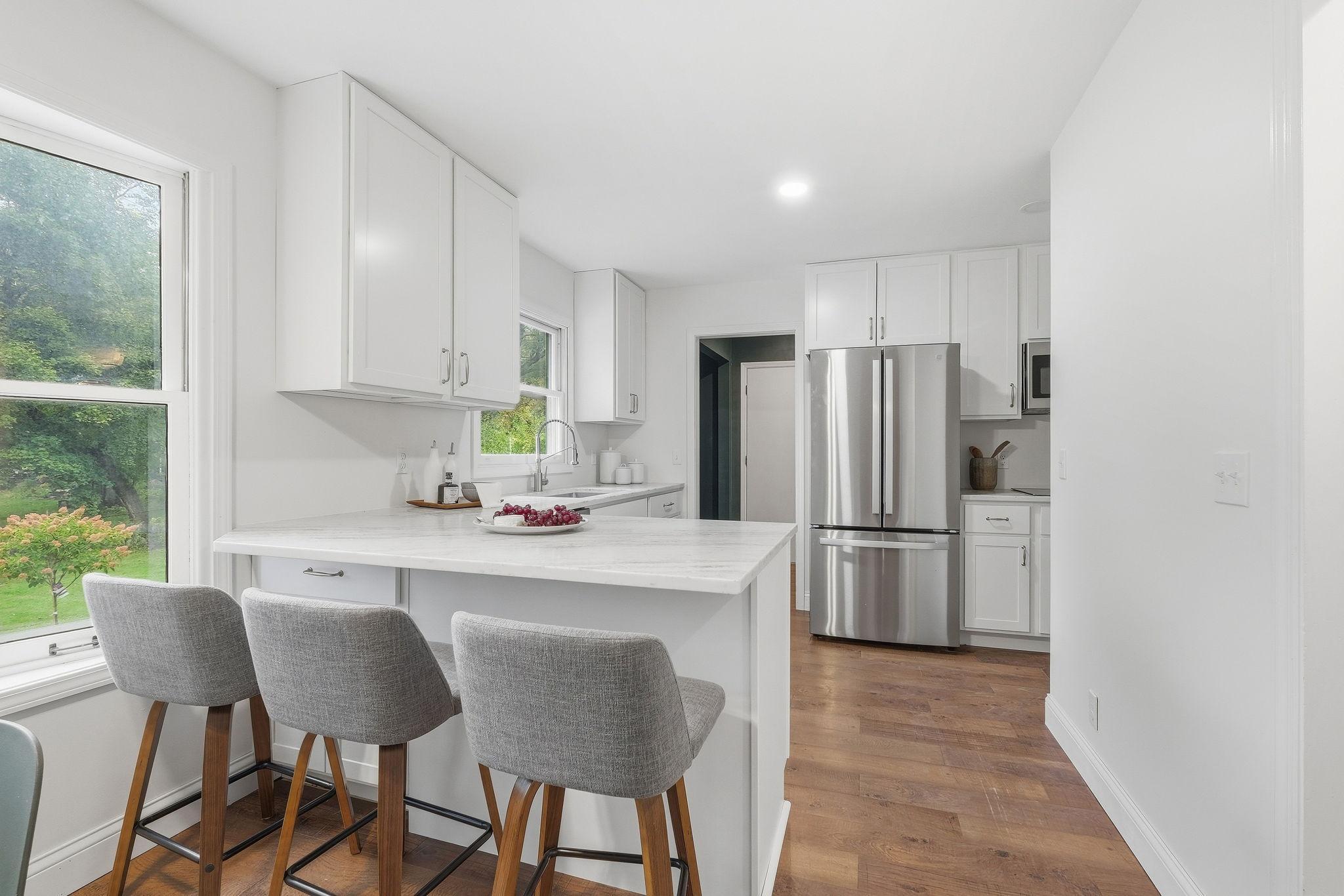 Bright white kitchen with a large island, gray bar stools, stainless steel appliances, and wood flooring.
