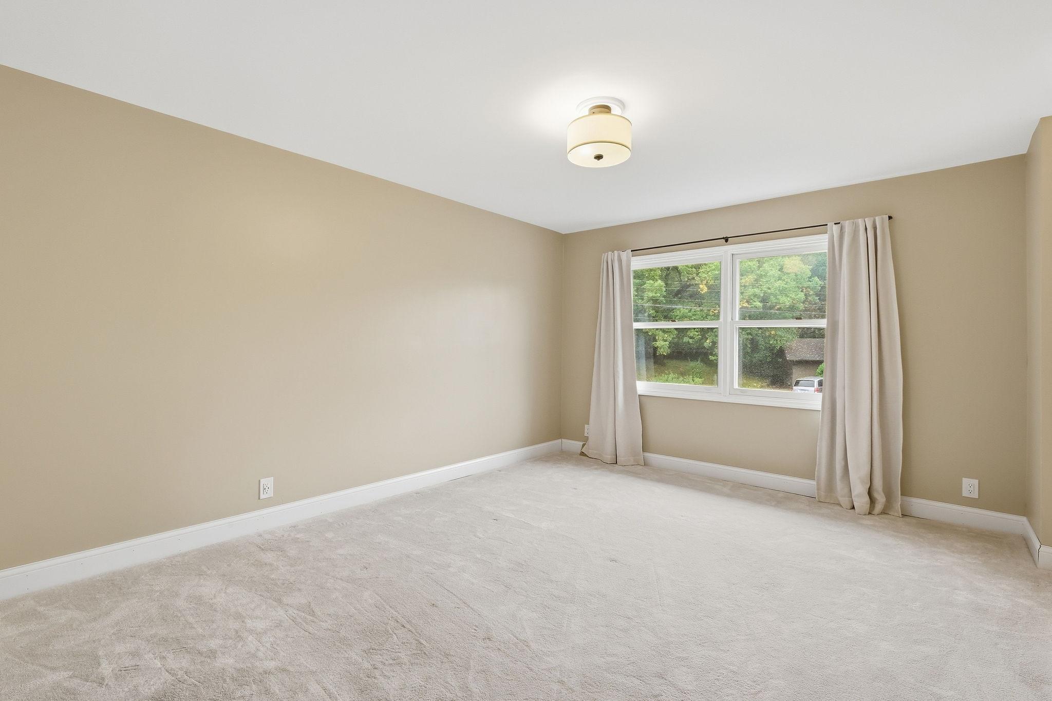 Empty bedroom with light carpet and a curtained window overlooking trees.