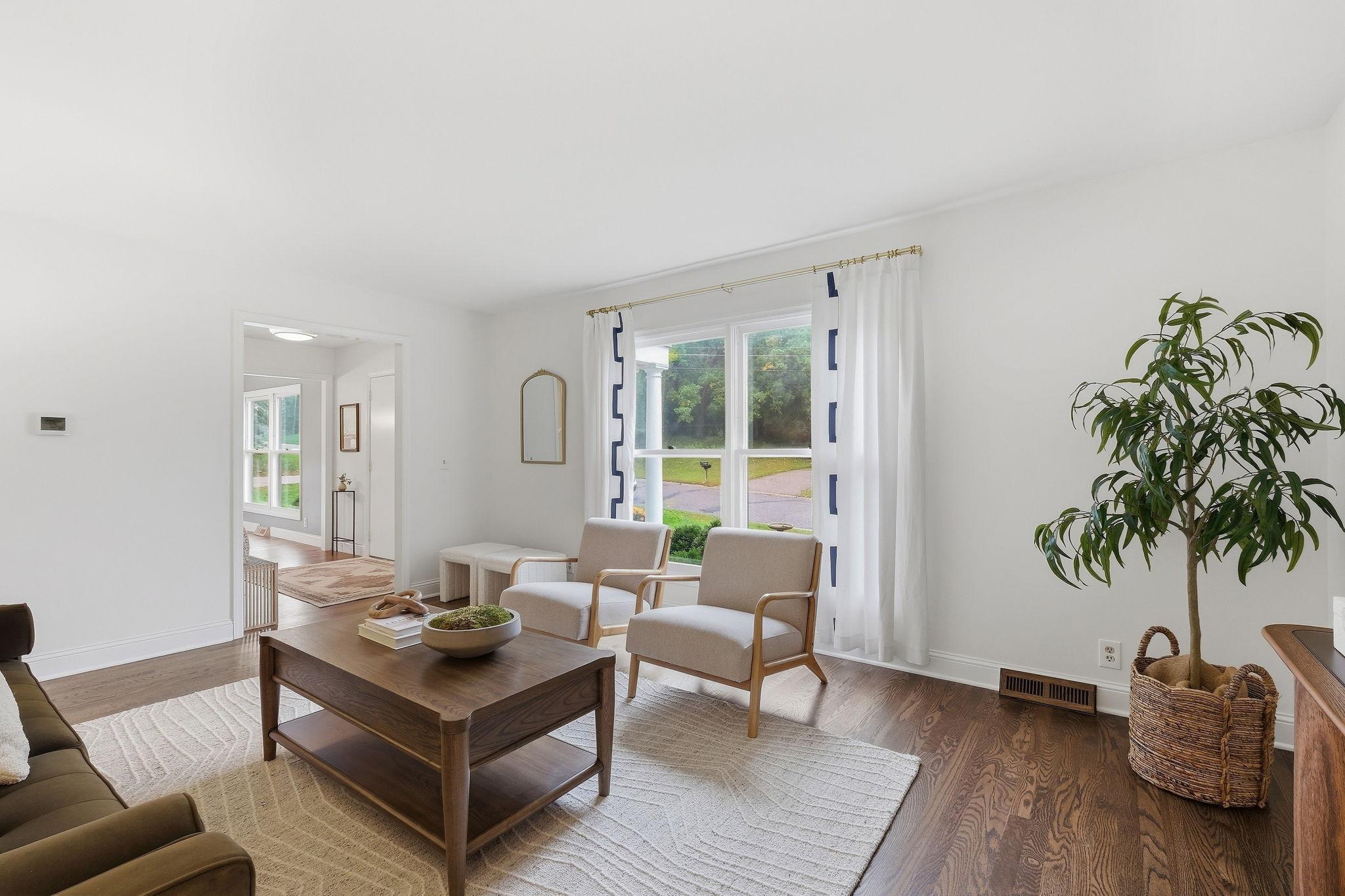 Staged living room with two light chairs, a dark wood coffee table, and a large curtained window.