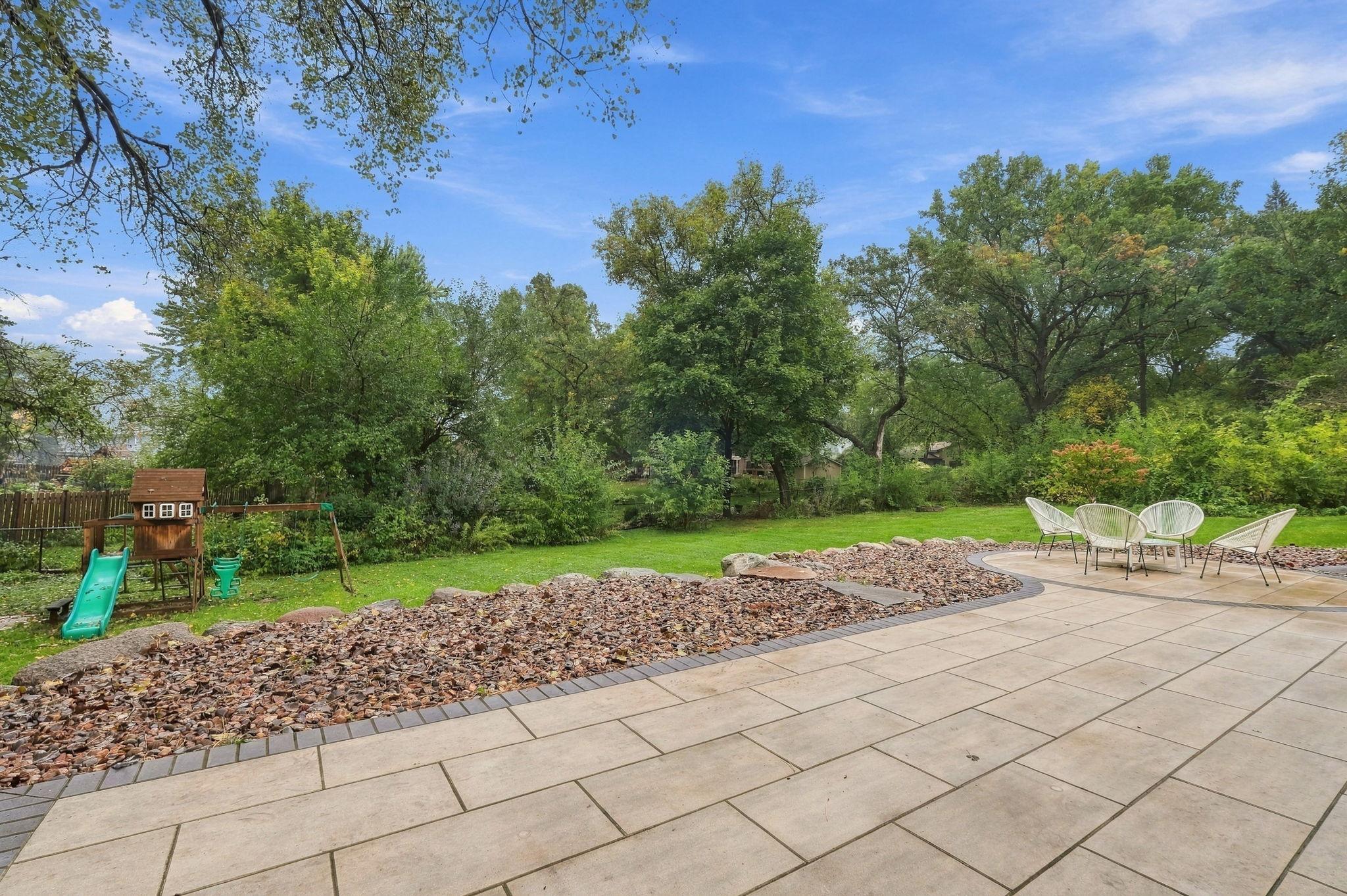 Backyard patio with stone pavers, white chairs, a children’s playset, and mature trees.