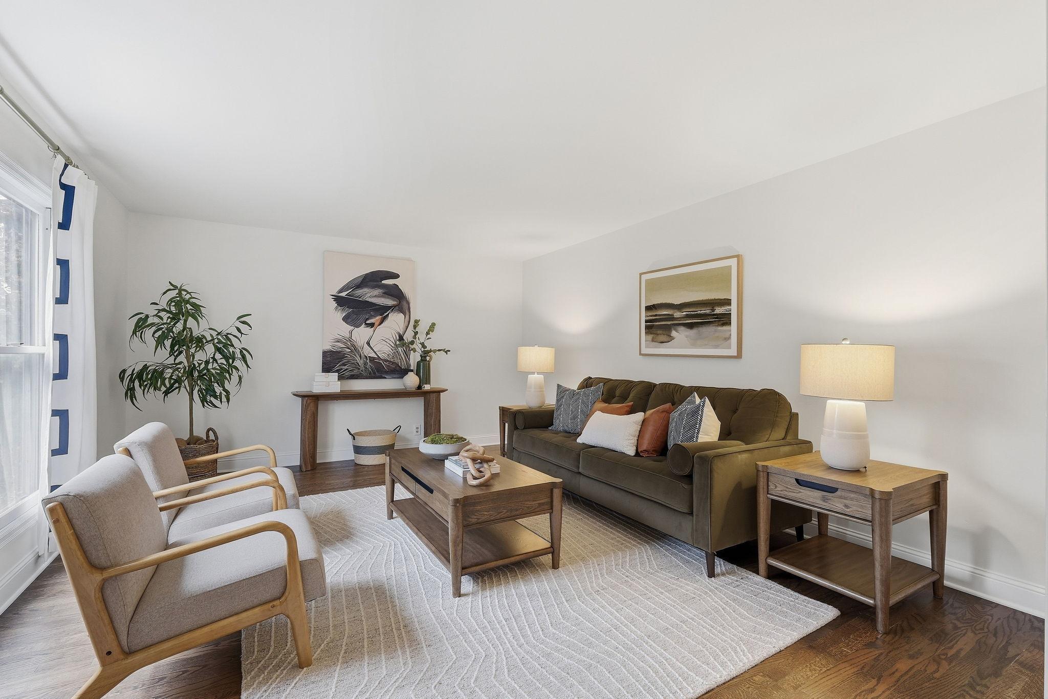 Staged living room with an olive sofa, wooden coffee table, two armchairs, and a large window.
