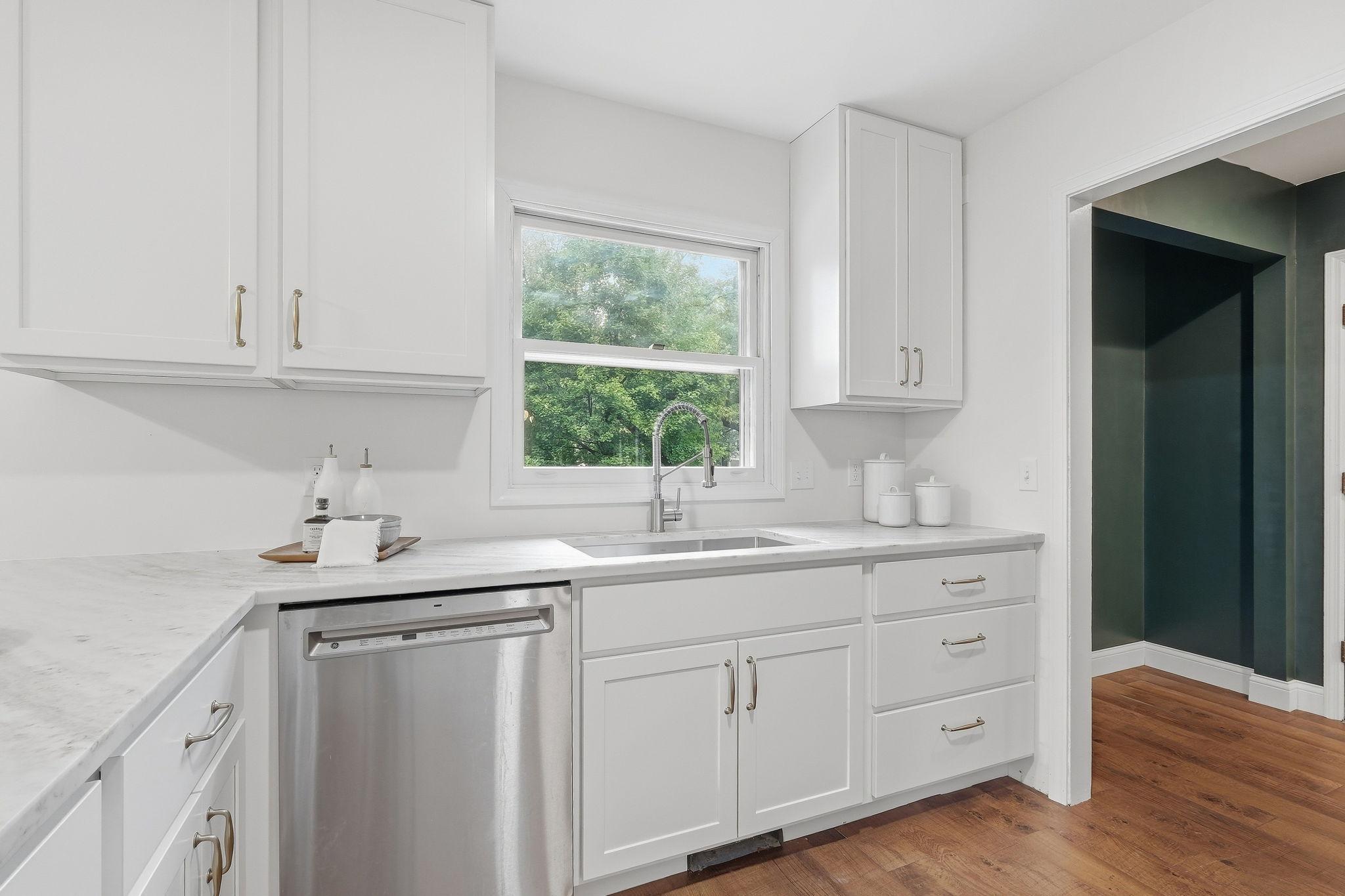 Bright white kitchen with pale stone counters, a stainless dishwasher, and a sink beneath a leafy window.