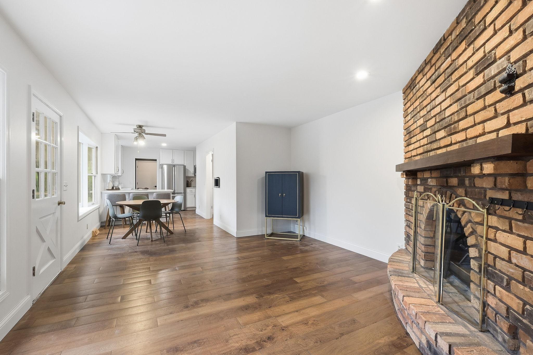 Open-plan living and dining area with a brick fireplace and wood floors; kitchen and dining table beyond.