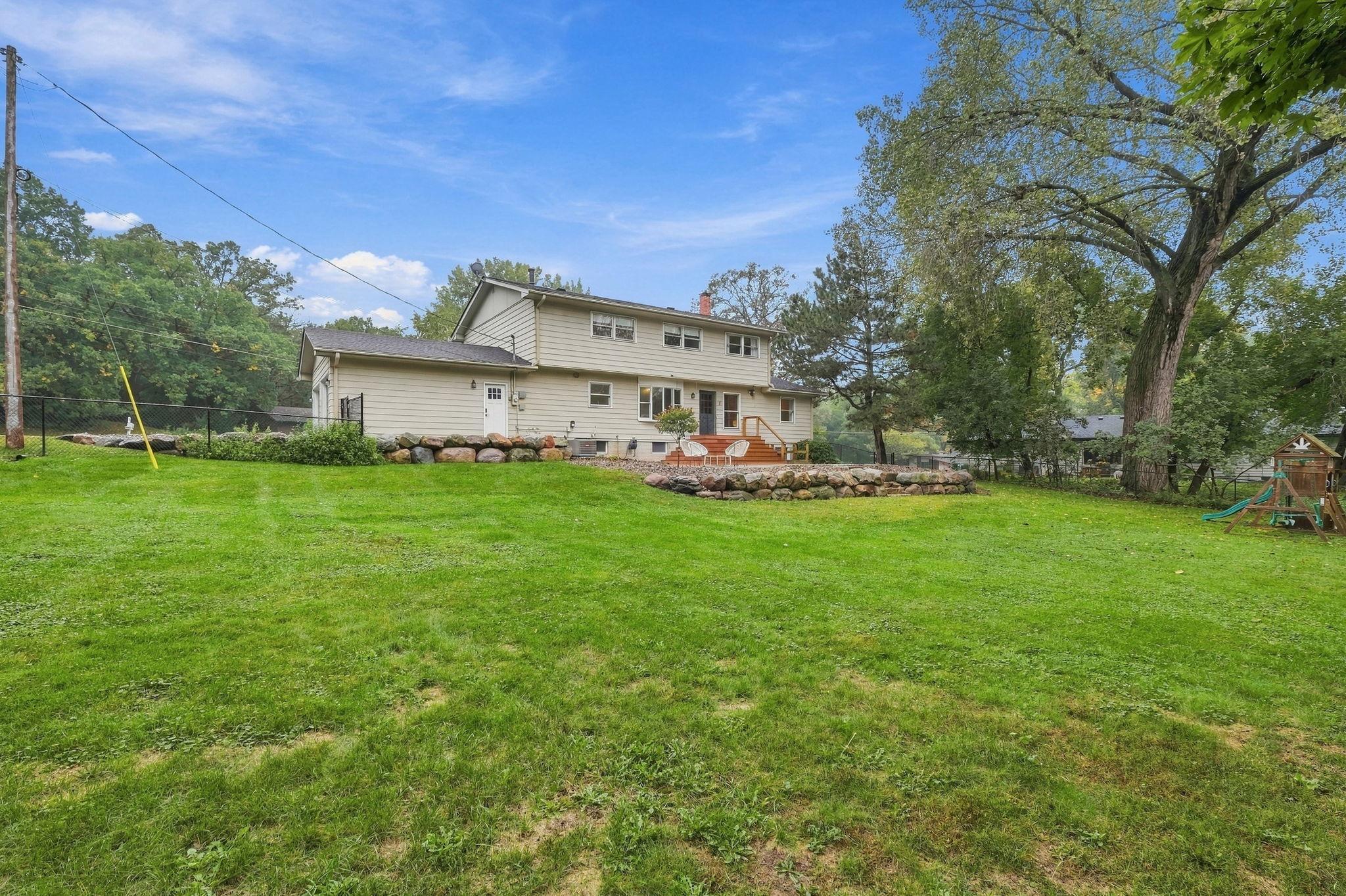 Rear of a beige two-story home with a raised deck, stone walls, and a broad lawn beneath mature trees.