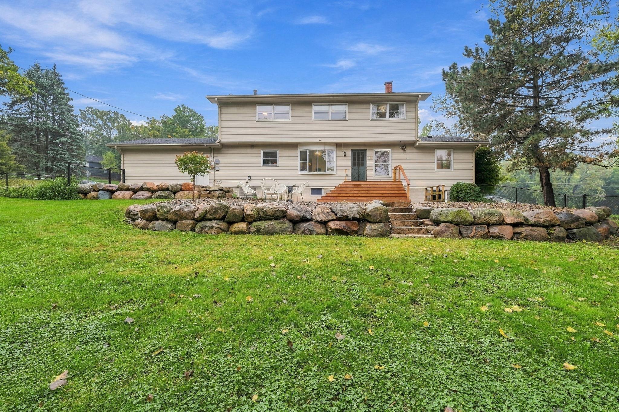 Rear of a two-story beige house with a wooden deck, stone retaining walls, and a broad lawn.