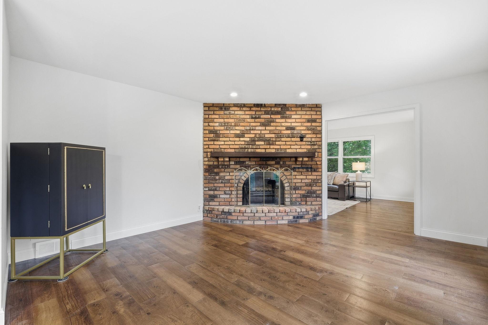 Living room with a curved brick fireplace, dark wood floors, and a doorway to a furnished sitting area.
