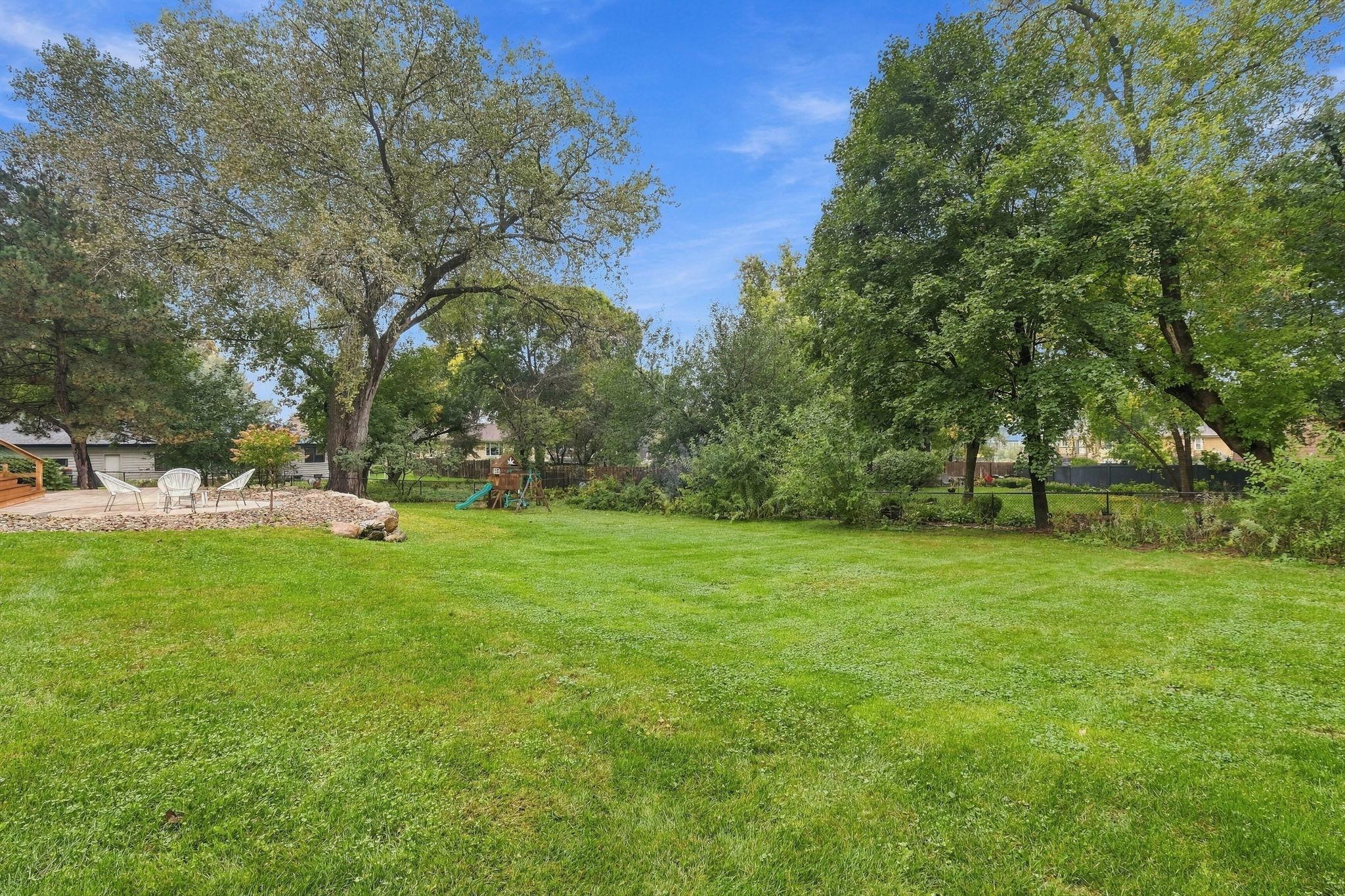 Spacious backyard with a broad lawn, mature trees, patio seating, and a children's playset.